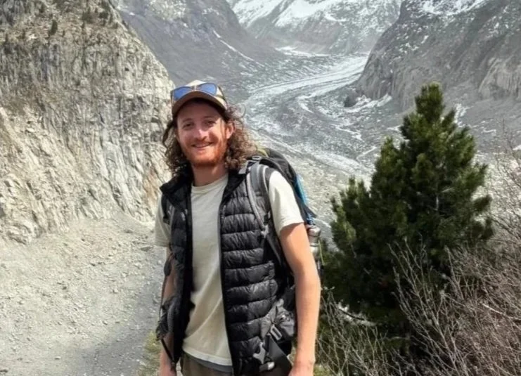 Aimsinlove client young man smiling outdoors in a mountainous area with rocky cliffs, a glacier or snow-covered mountain, and a small green tree.