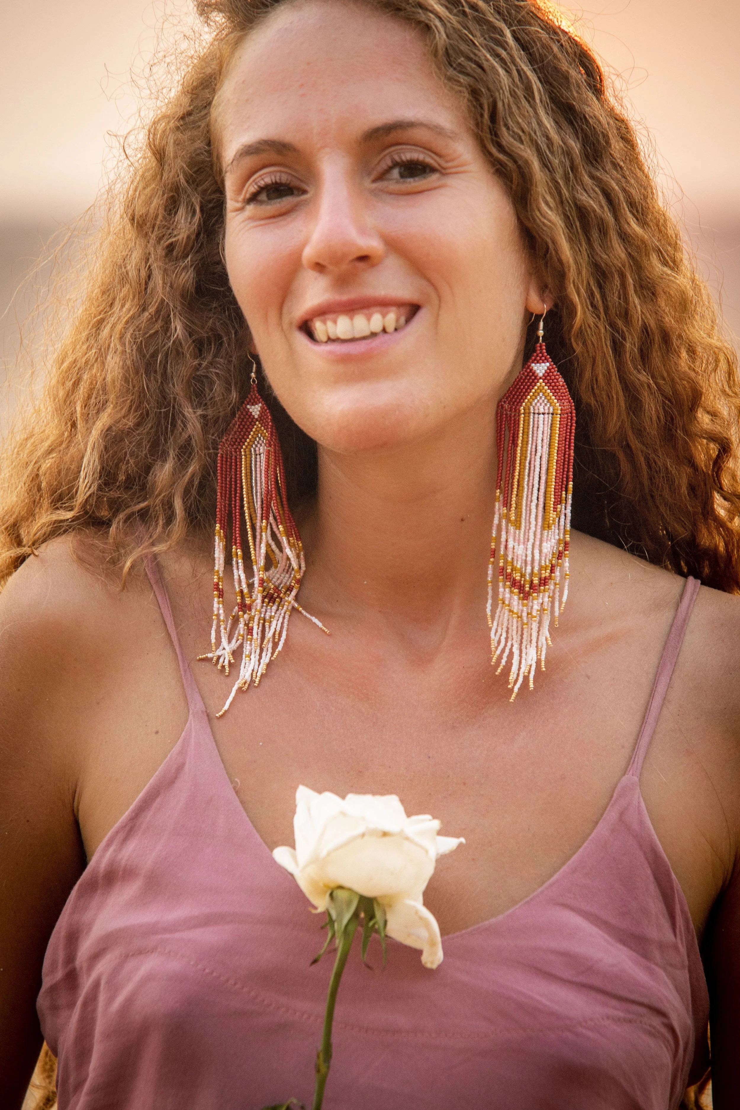 A woman with curly red hair wearing large beaded earrings and a pink spaghetti strap top holding a white flower. She is smiling and appears to be outdoors near sunset.