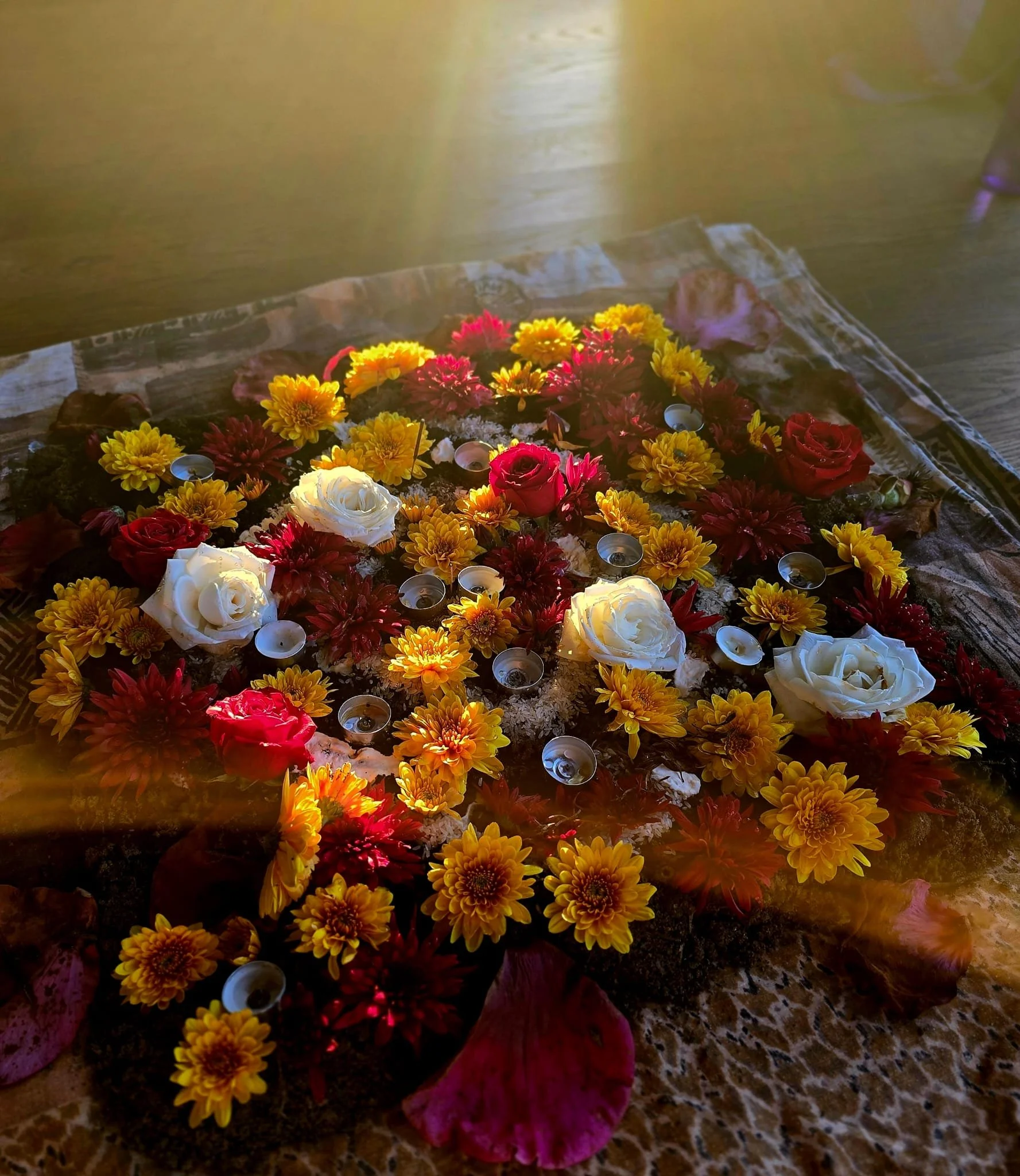 Decorative flower arrangement on a wooden table, featuring yellow, red, and white flowers, surrounded by small tea light candles and red leaves, with a warm golden background.