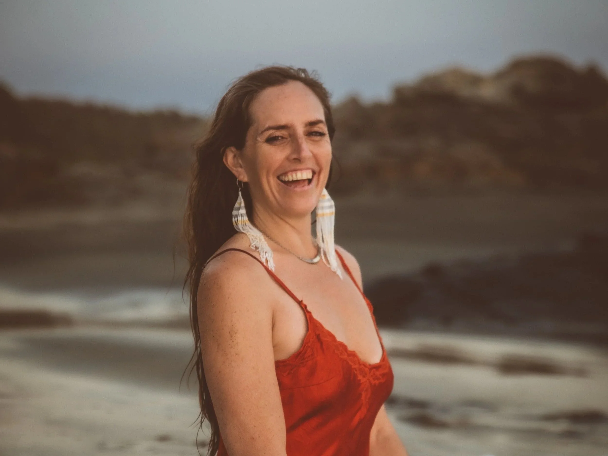 A woman with long brown hair smiling at the beach, wearing a red lace dress with spaghetti straps.
