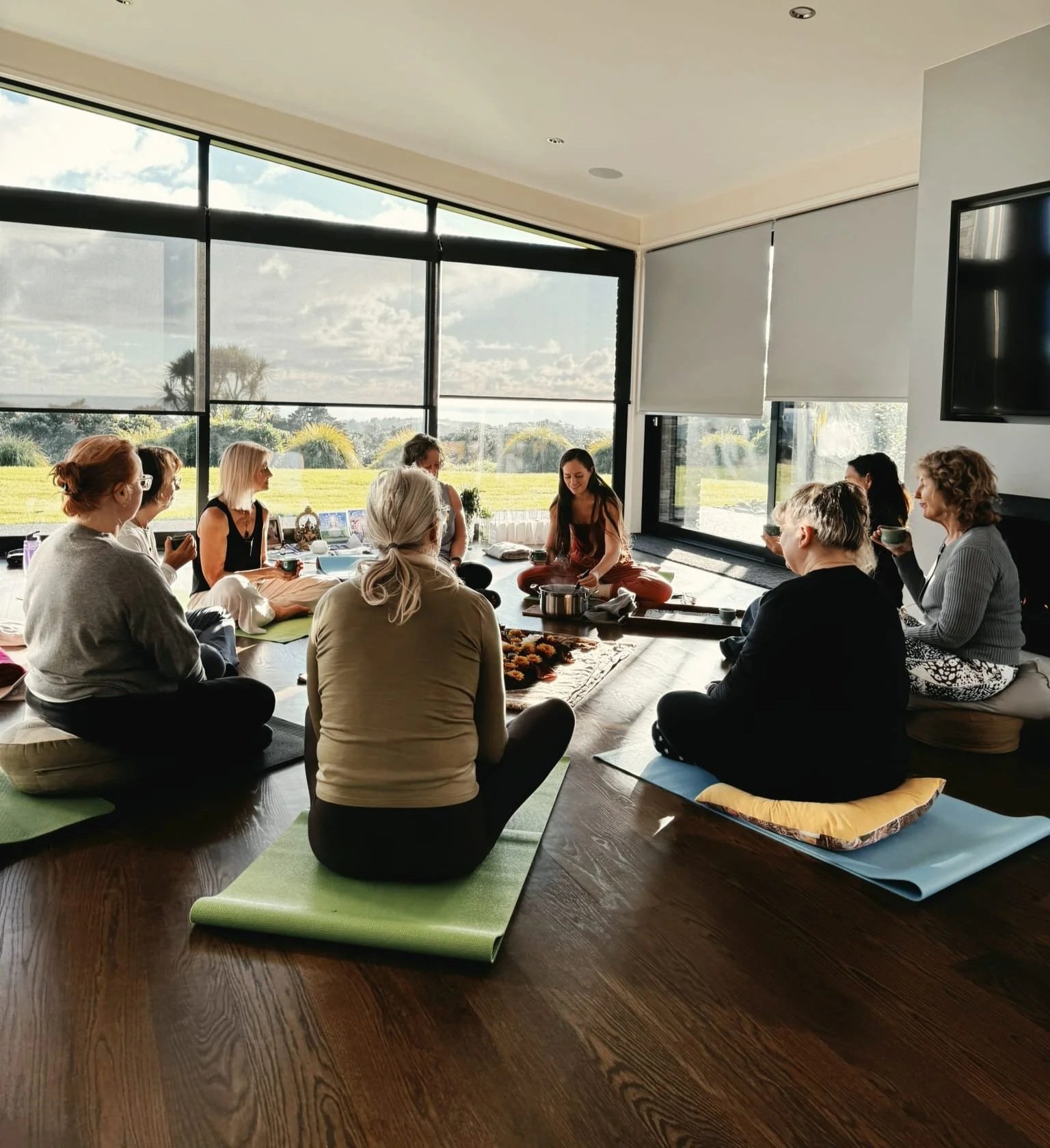 Group of people participating in a meditation or yoga session indoors on mats, with large windows showing a green landscape outside.