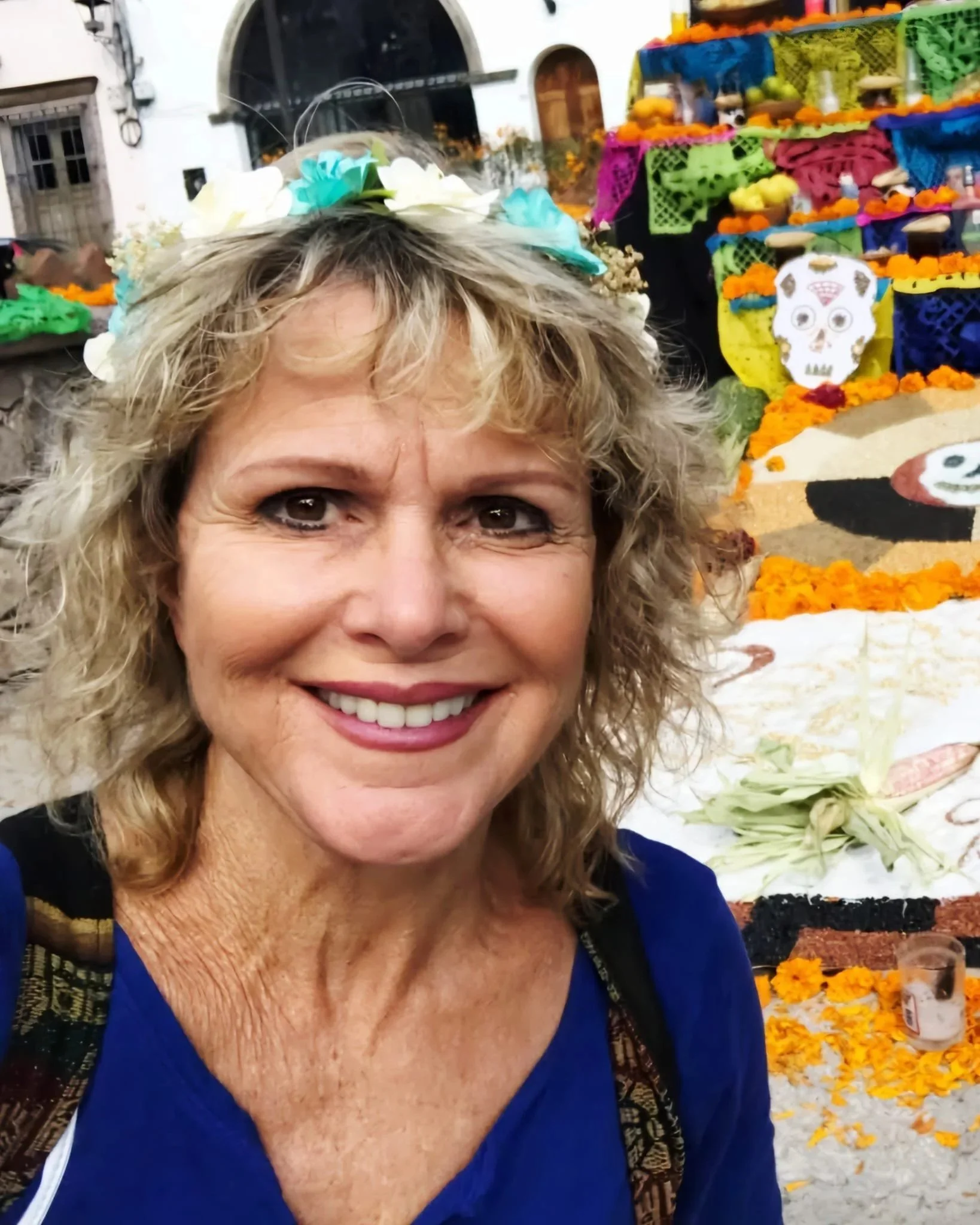 A smiling woman with curly blonde hair wearing a blue top, standing in front of a colorful decorated altar with marigold flowers, skull motifs, and traditional Mexican decorations.