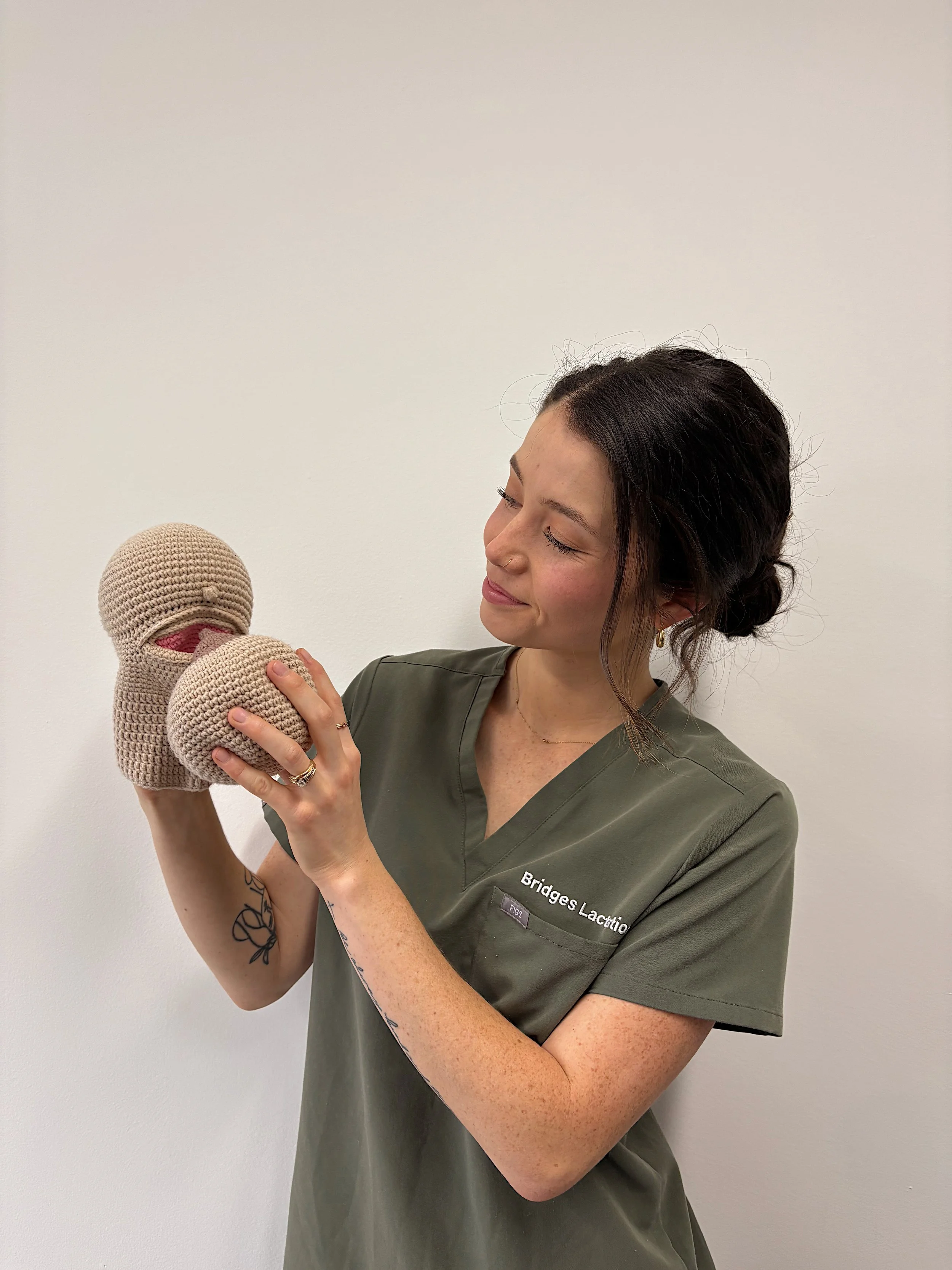 A woman in medical scrubs holding a crochet toy in the shape of a breast, smiling while looking at it.