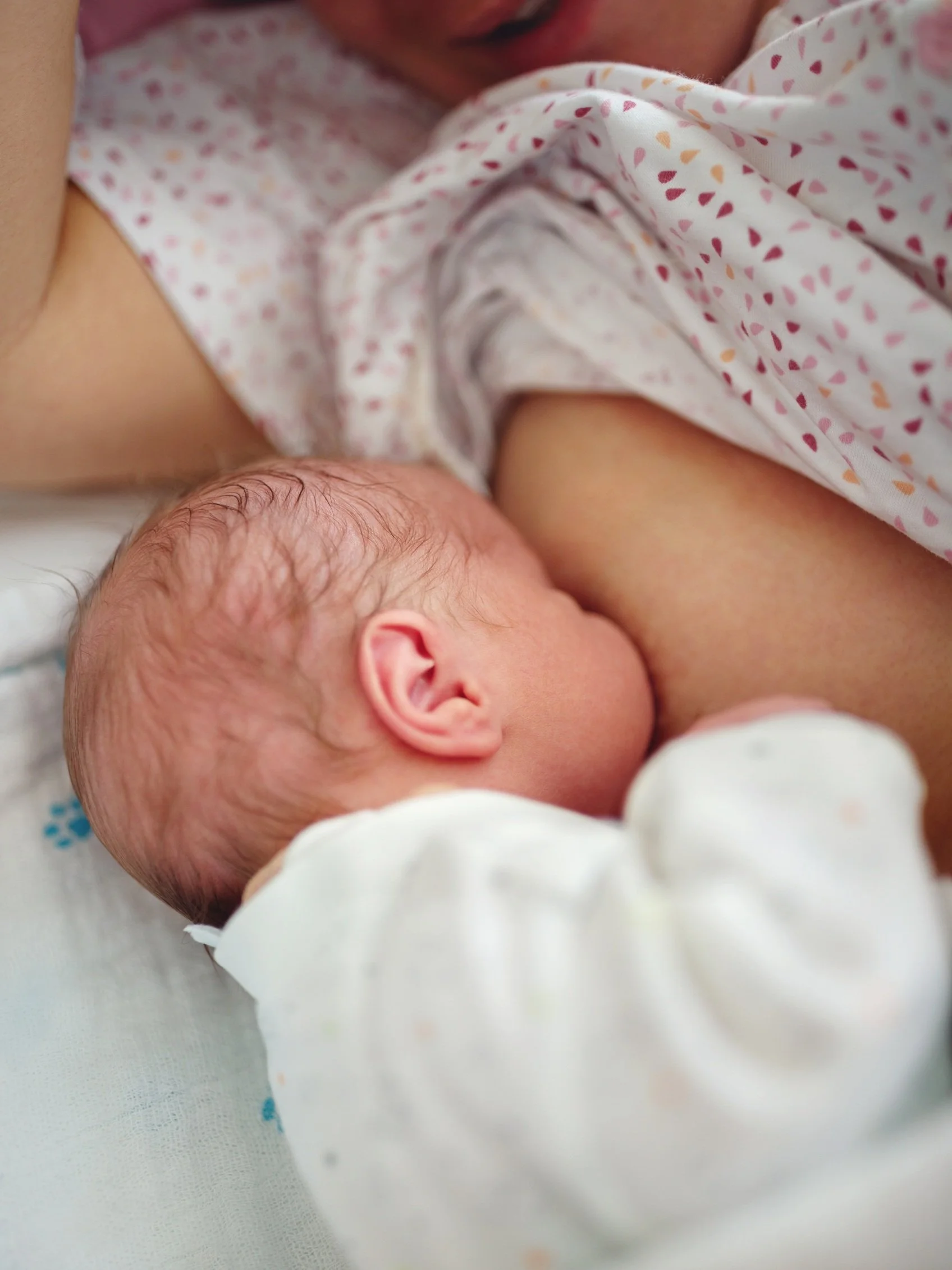 A newborn baby breastfeeding, lying on their side with their head near their mother’s breast, wearing a white onesie and a blanket with pink, orange, and purple hearts.