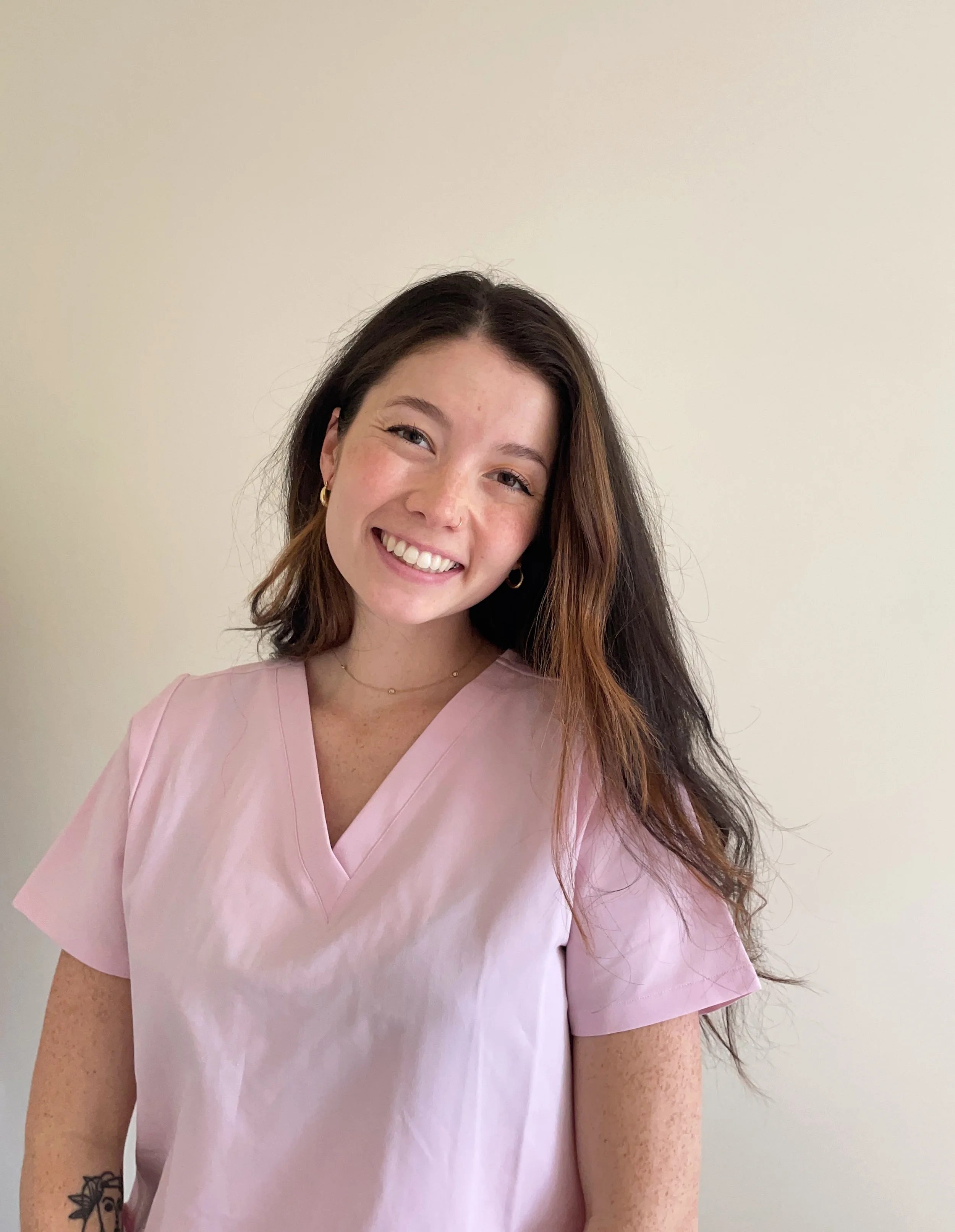 Young woman with long brown hair, smiling, wearing pink medical scrubs, standing against a plain light-colored wall.