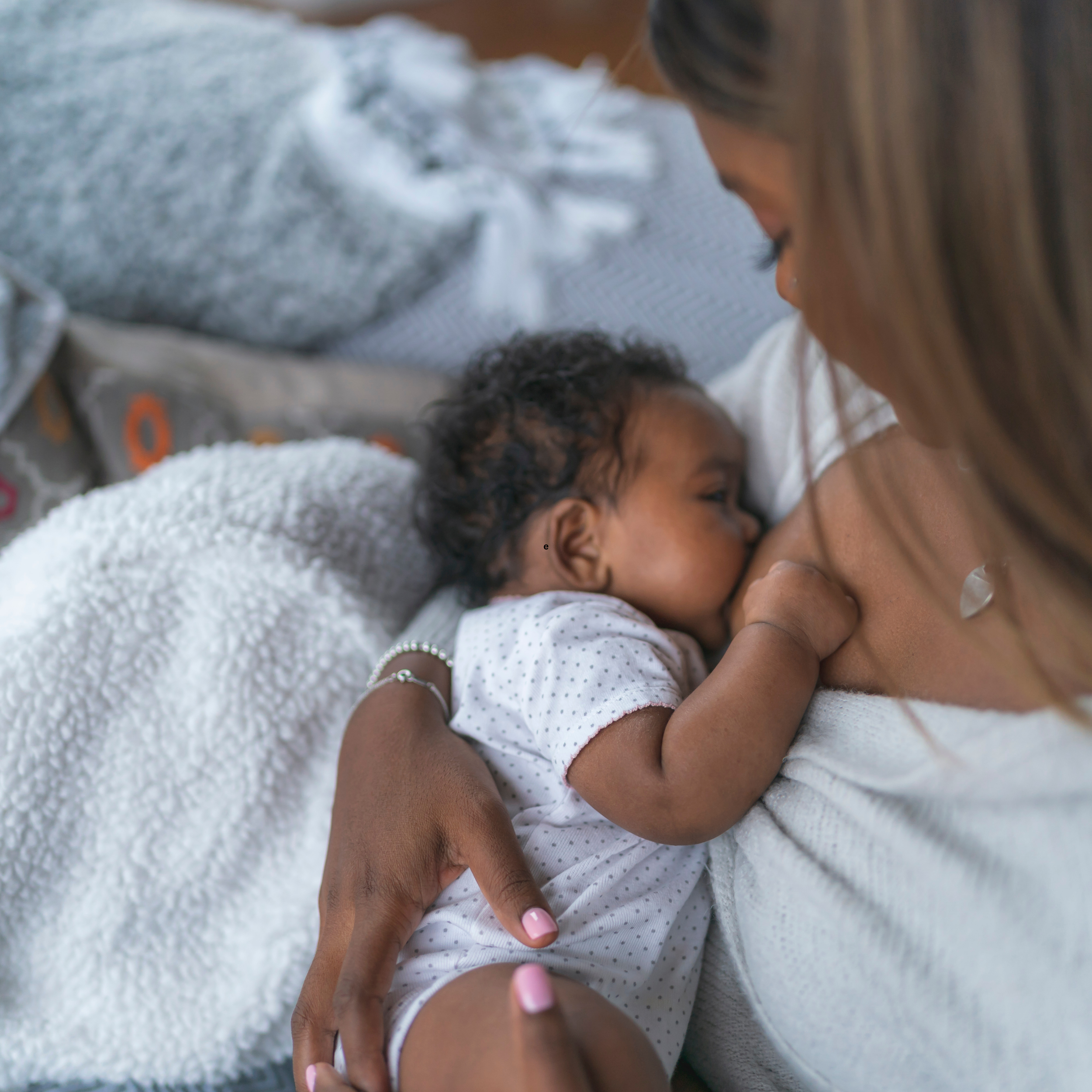 A mother breastfeeding her infant, lying on a bed wrapped in a white blanket.