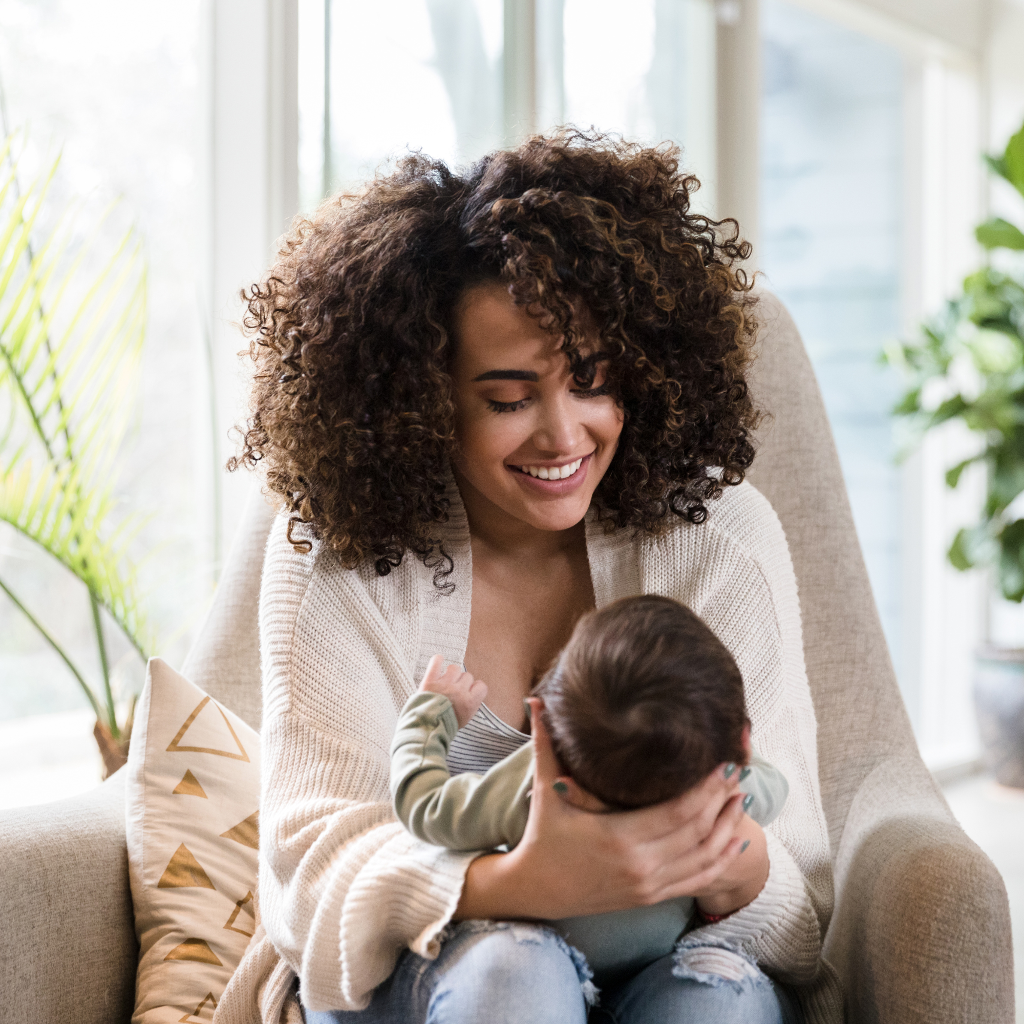 A woman holding a baby on a beige couch in a bright living room with large windows and green plants.