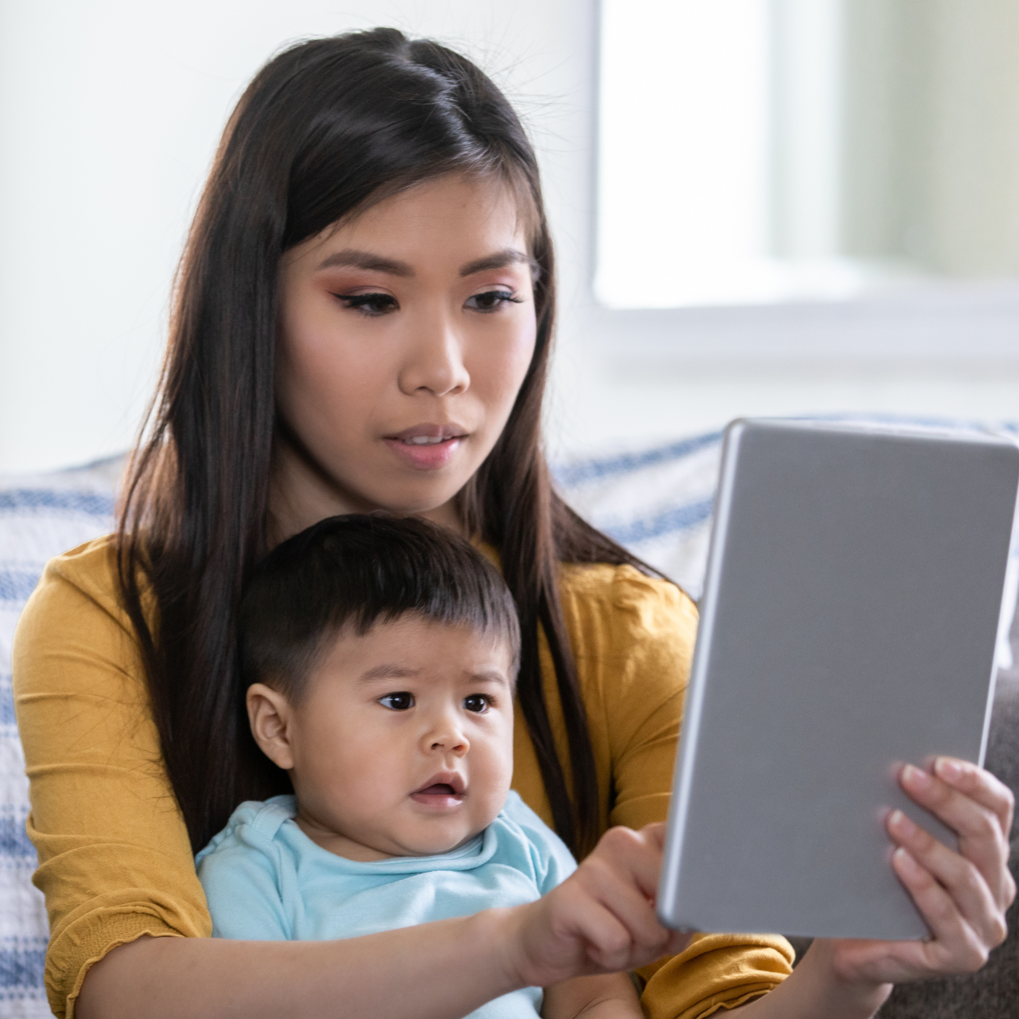 A woman and a young boy sitting on a couch looking at a tablet, in a well-lit room.
