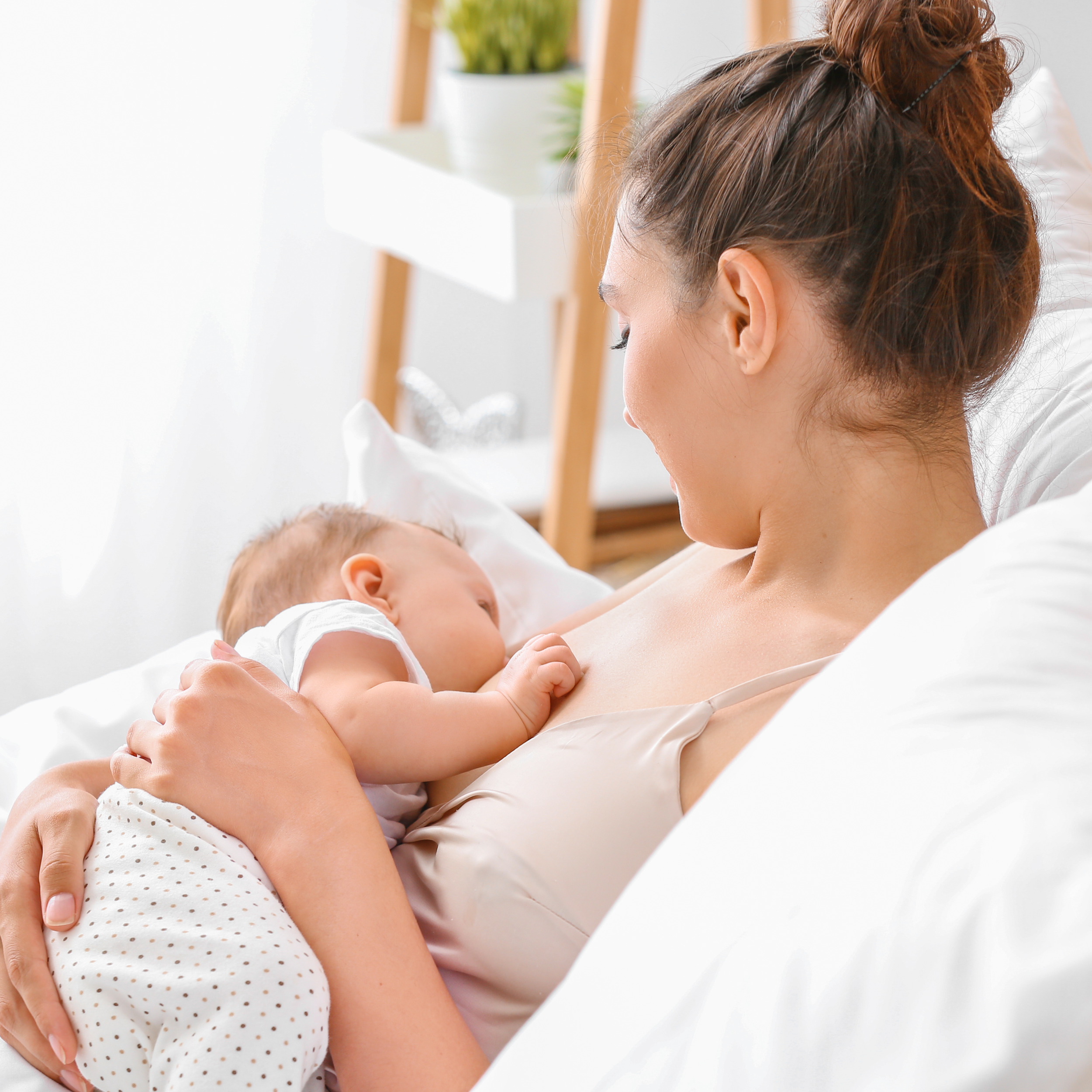 A woman breastfeeding a baby in a bright, cozy room with a plant and bookshelf in the background.