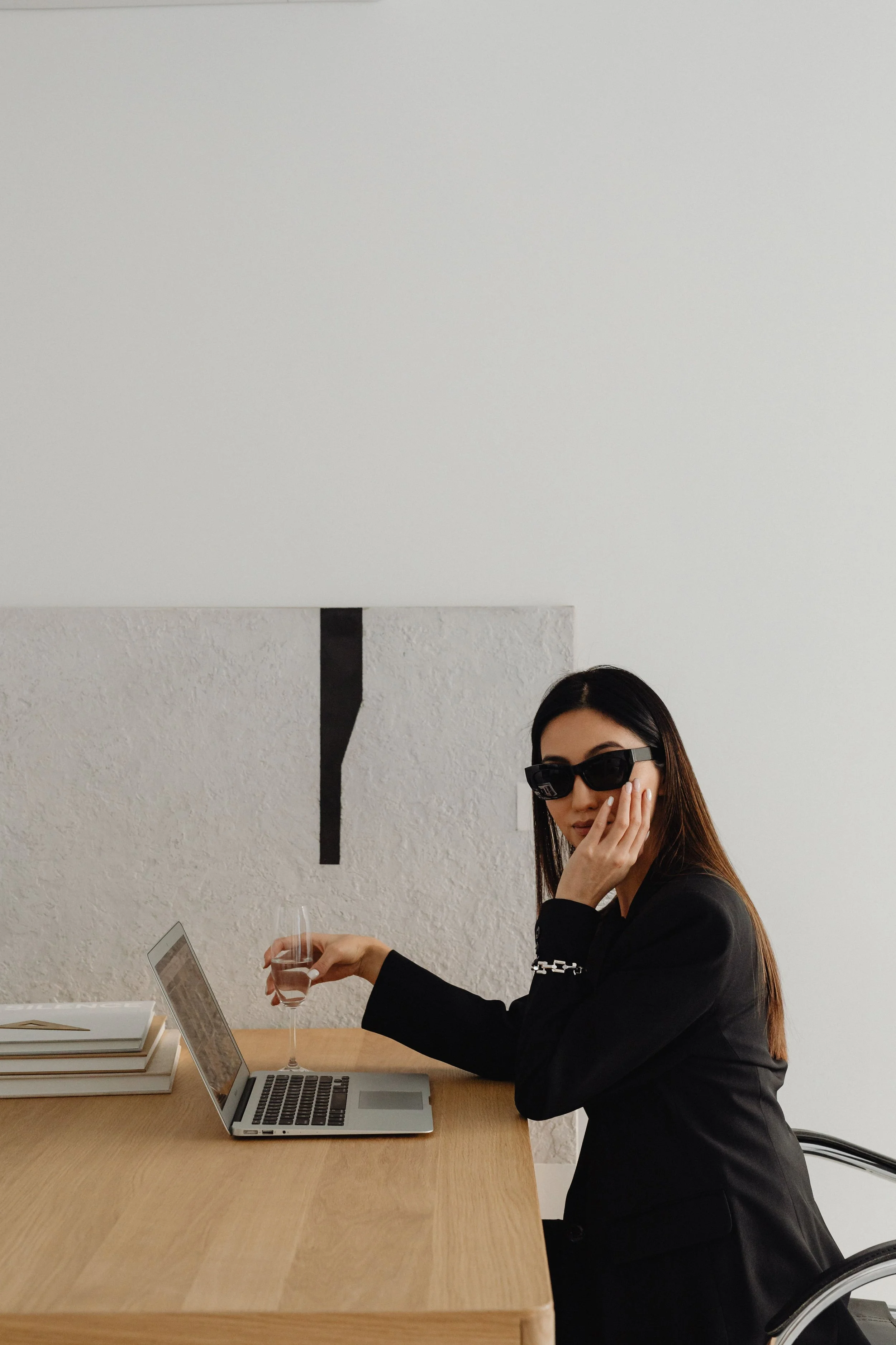 A woman sitting at a desk with a laptop, holding a glass of water, wearing sunglasses and a black blazer, with a minimalist white wall background.