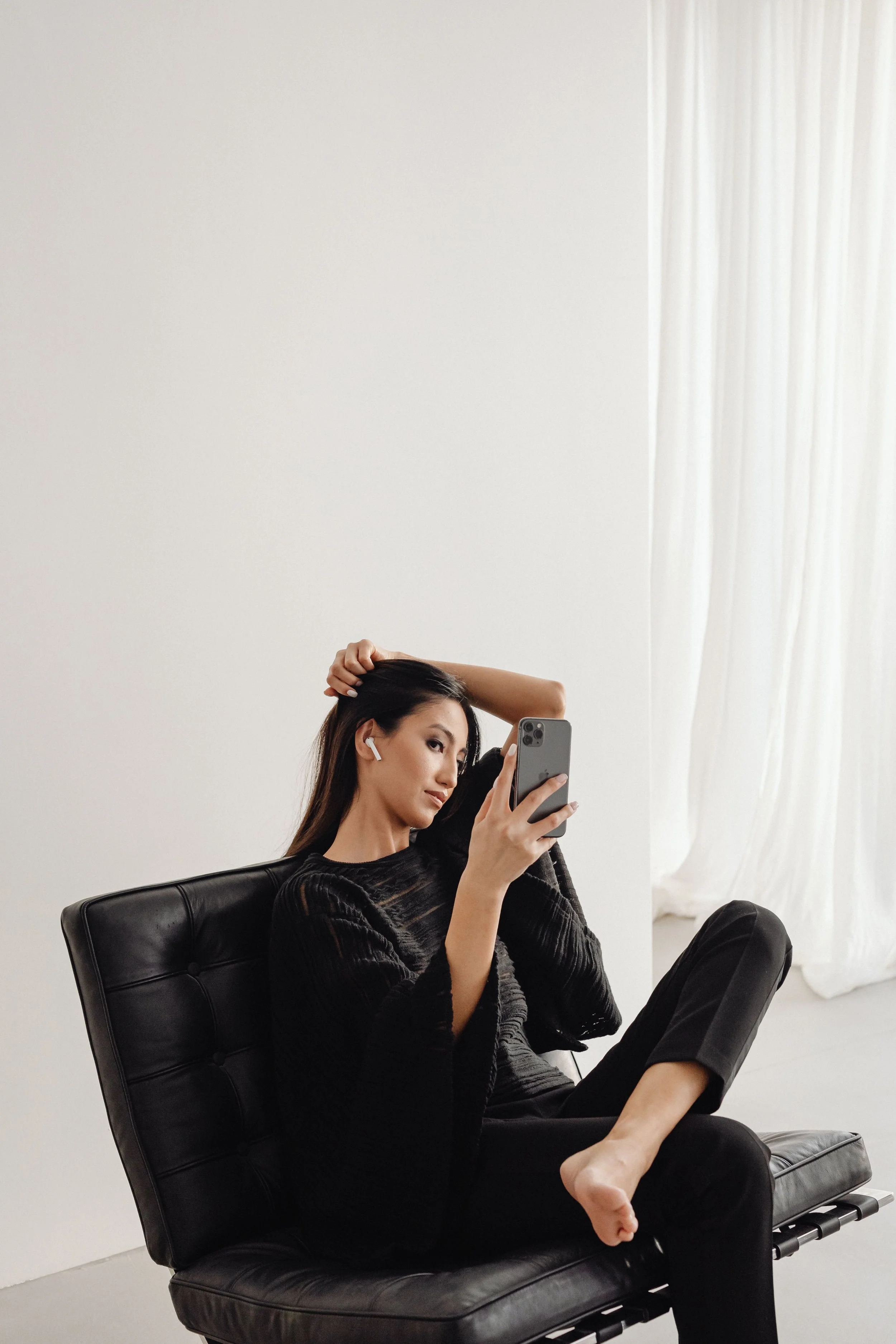 A woman sitting on a black leather chair takes a selfie with her smartphone. She is wearing black earphones and a black top, with white curtains and a white wall in the background.