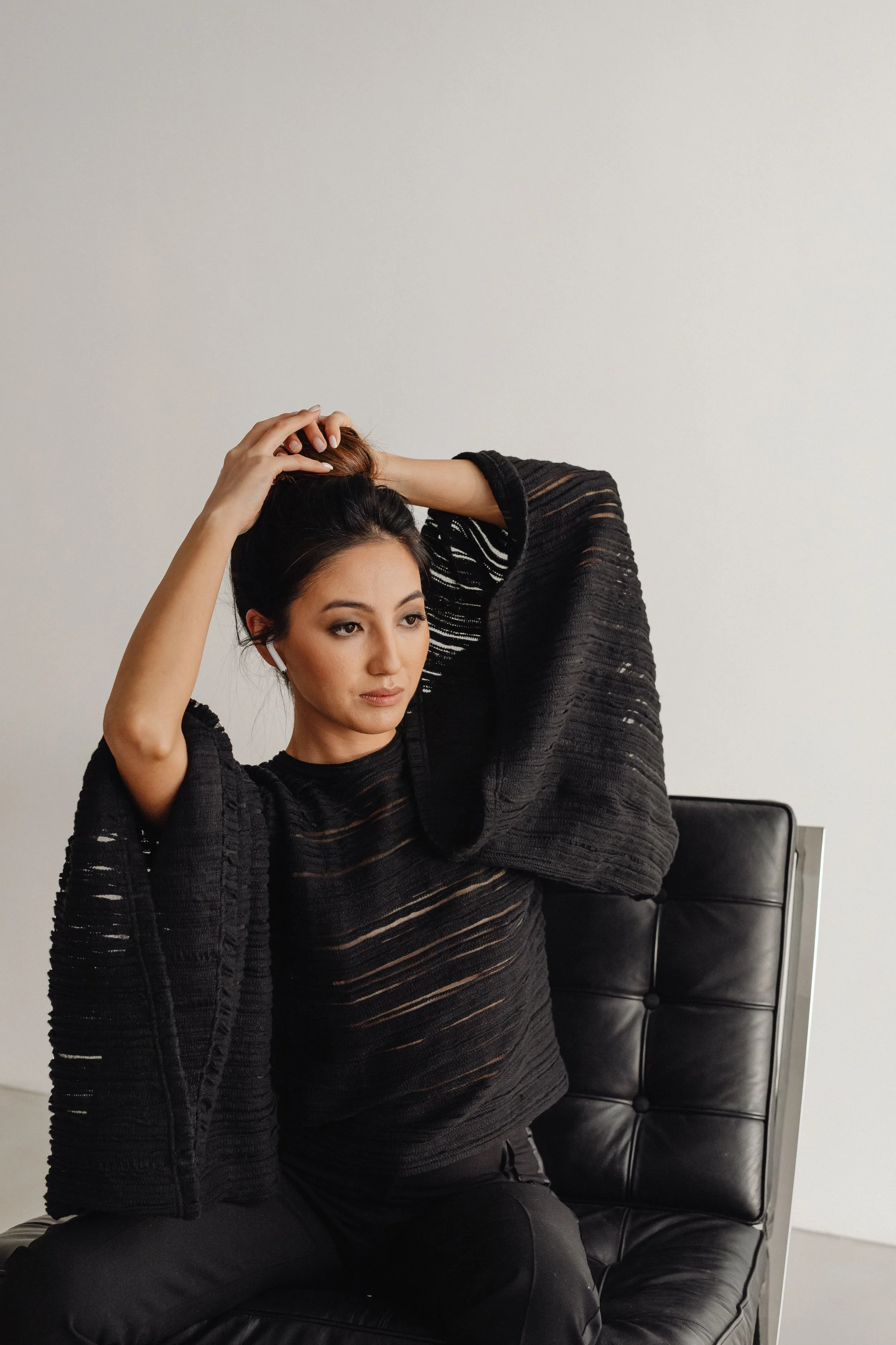 A woman sitting on a black leather chair, wearing a black, semi-sheer, textured top, and adjusting her hair.