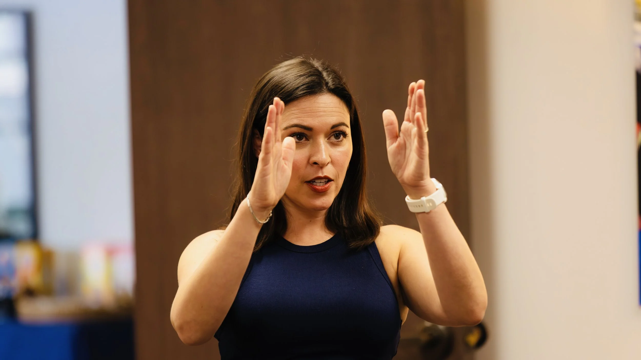 A woman with dark brown hair wearing a sleeveless navy blue top gestures with both hands near her face, appearing to be speaking or explaining something.