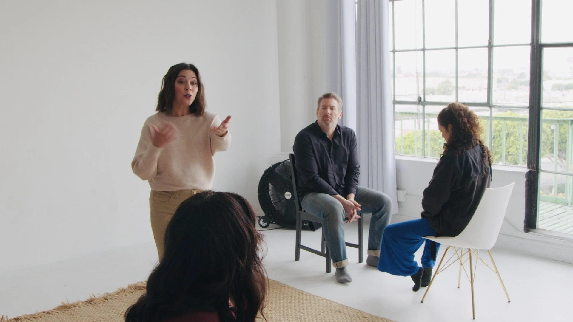 A woman with shoulder-length dark hair stands and speaks to three seated individuals in a bright, sparsely decorated room with large windows and white walls.