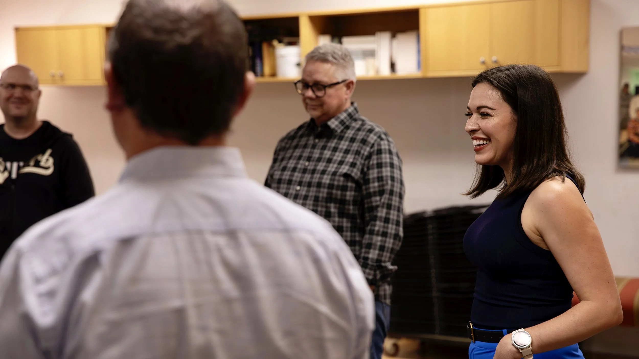 Group of four people talking in a casual office or community space, with a woman in a navy dress smiling and engaging in conversation.