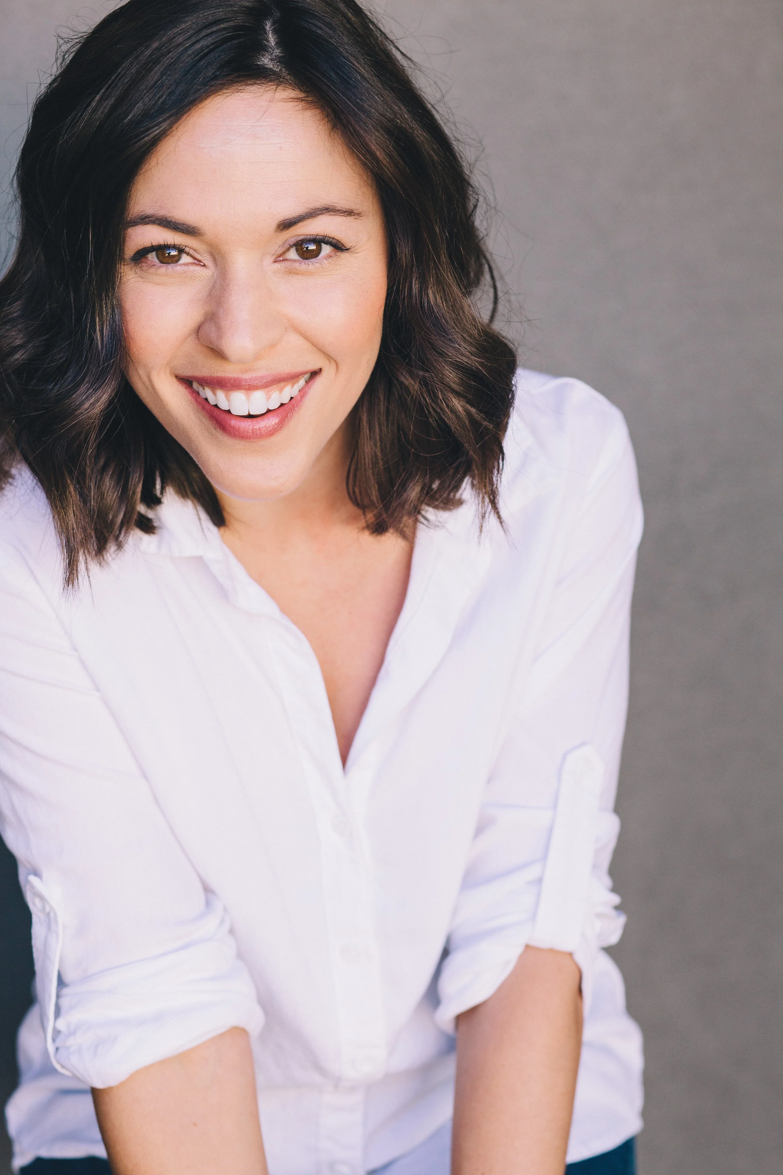 Close-up of a smiling woman with shoulder-length dark brown hair, wearing a white button-up shirt with rolled-up sleeves, against a gray background.