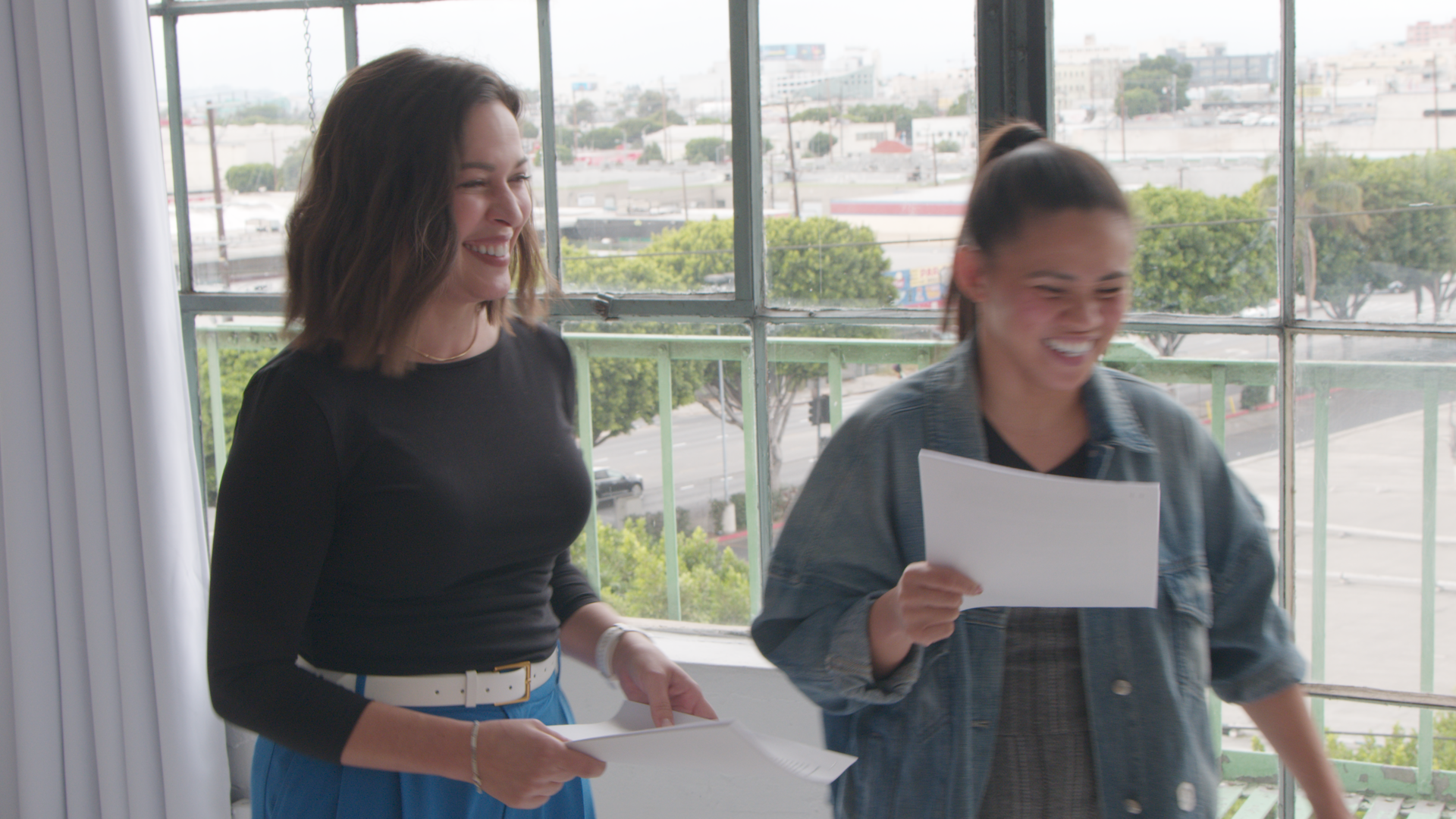 Two women, one with shoulder-length brown hair wearing a black top and blue pants, and the other with dark hair in a ponytail wearing a denim jacket, are smiling and holding papers, standing near a large window with cityscape views.