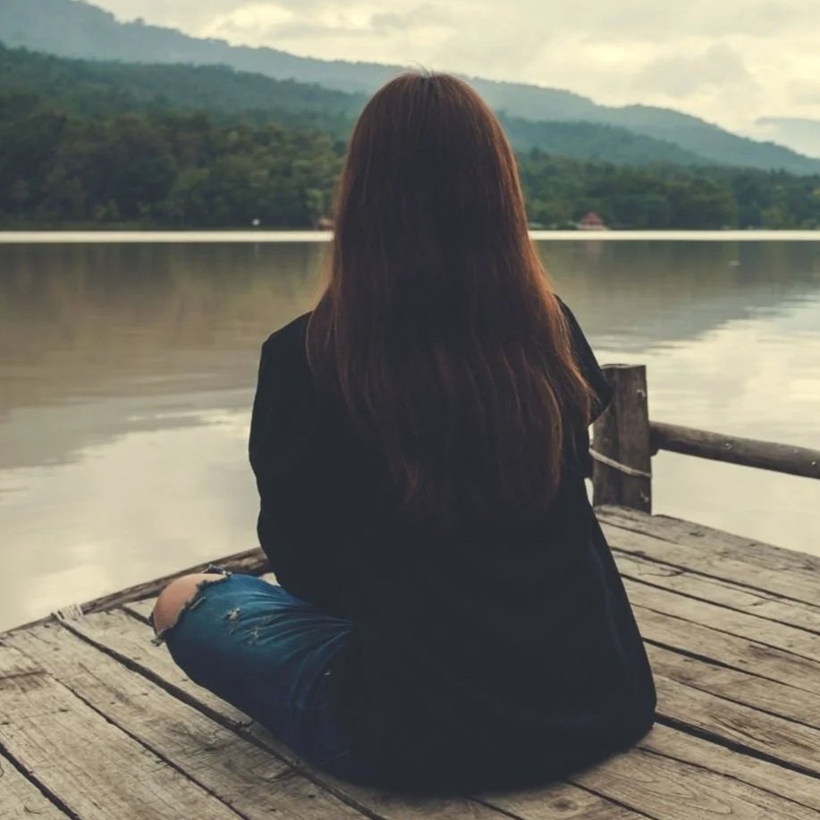 A woman with long brown hair sitting cross-legged on a wooden dock, looking out over a calm lake with mountains and forest in the background during cloudy weather.