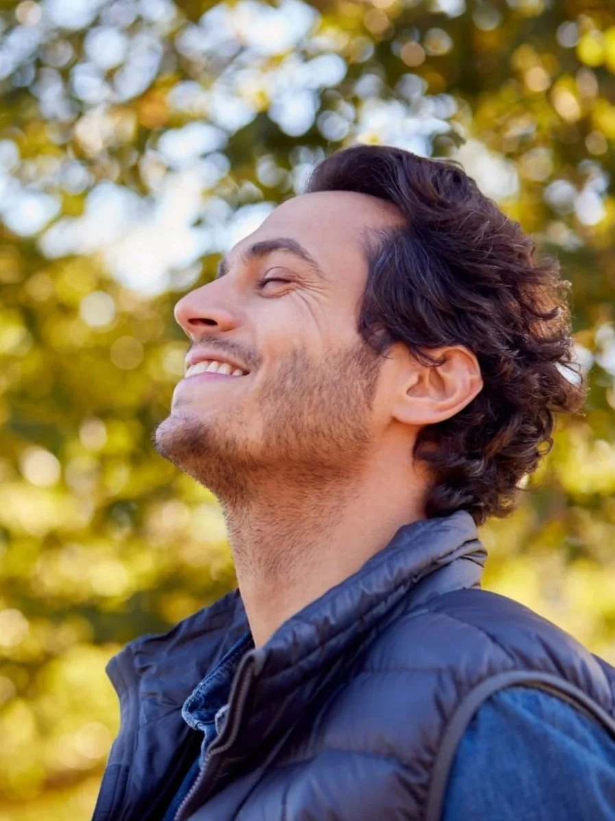 Side view of smiling man with eyes closed, outdoor background of blurred autumn leaves, wearing a dark jacket.
