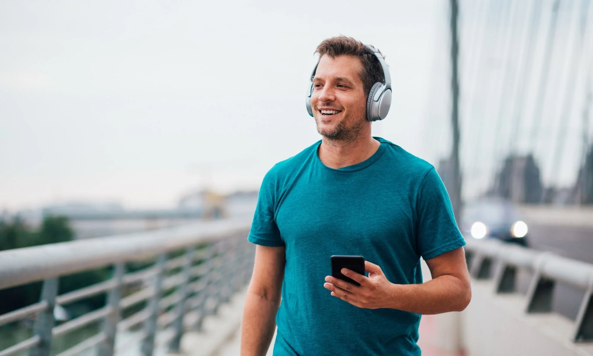 Man in blue T-shirt with headphones smiling while walking on bridge and holding smartphone.
