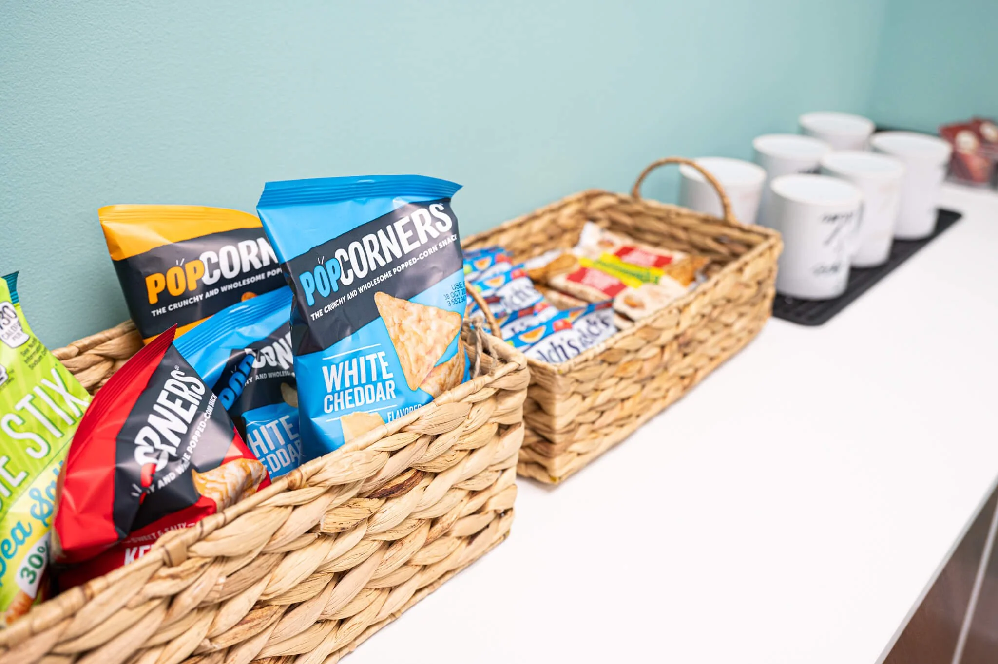 A picture of two rectangular woven baskets on the left there are bags of popcorners chips in the on on th right there are fruit snacks and snack bars. In the distance there are mugs, the surface is white and the wall is teal, therapist waiting rooom