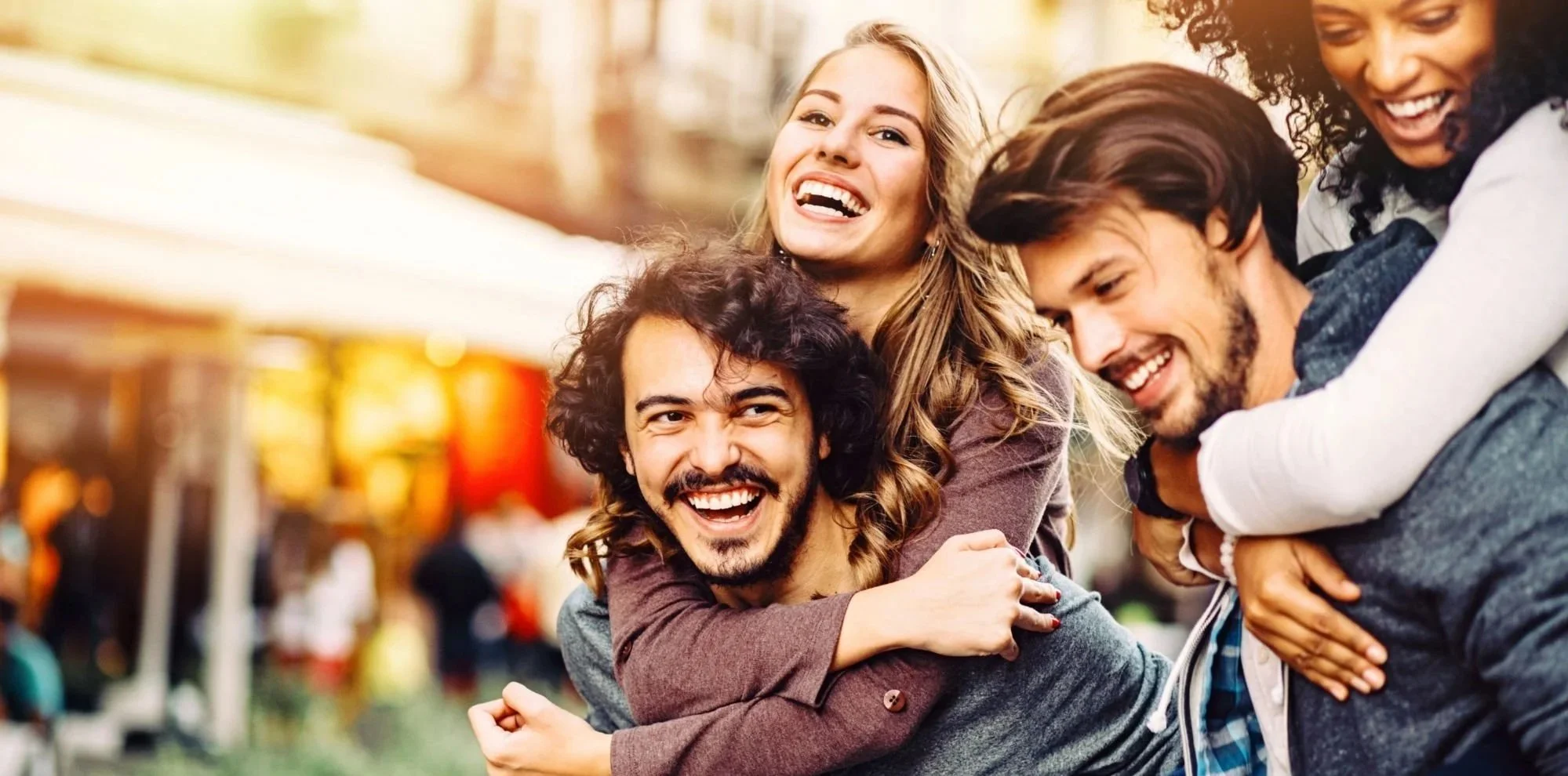Group of five young adults smiling and hugging at an outdoor fair or festival with colorful booths in the background.