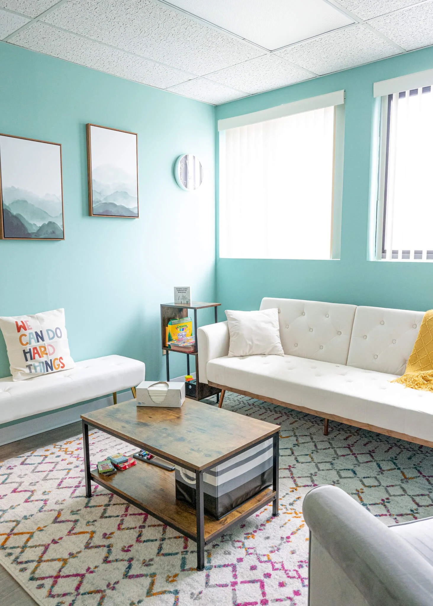An office with a white couch, a wooden coffee table with a tissue box, teal walls with two windows, two pictures on the wall of mountains. A white bench with a pillow saying we can do hard things. A therapist's office at Cope & Calm therapy