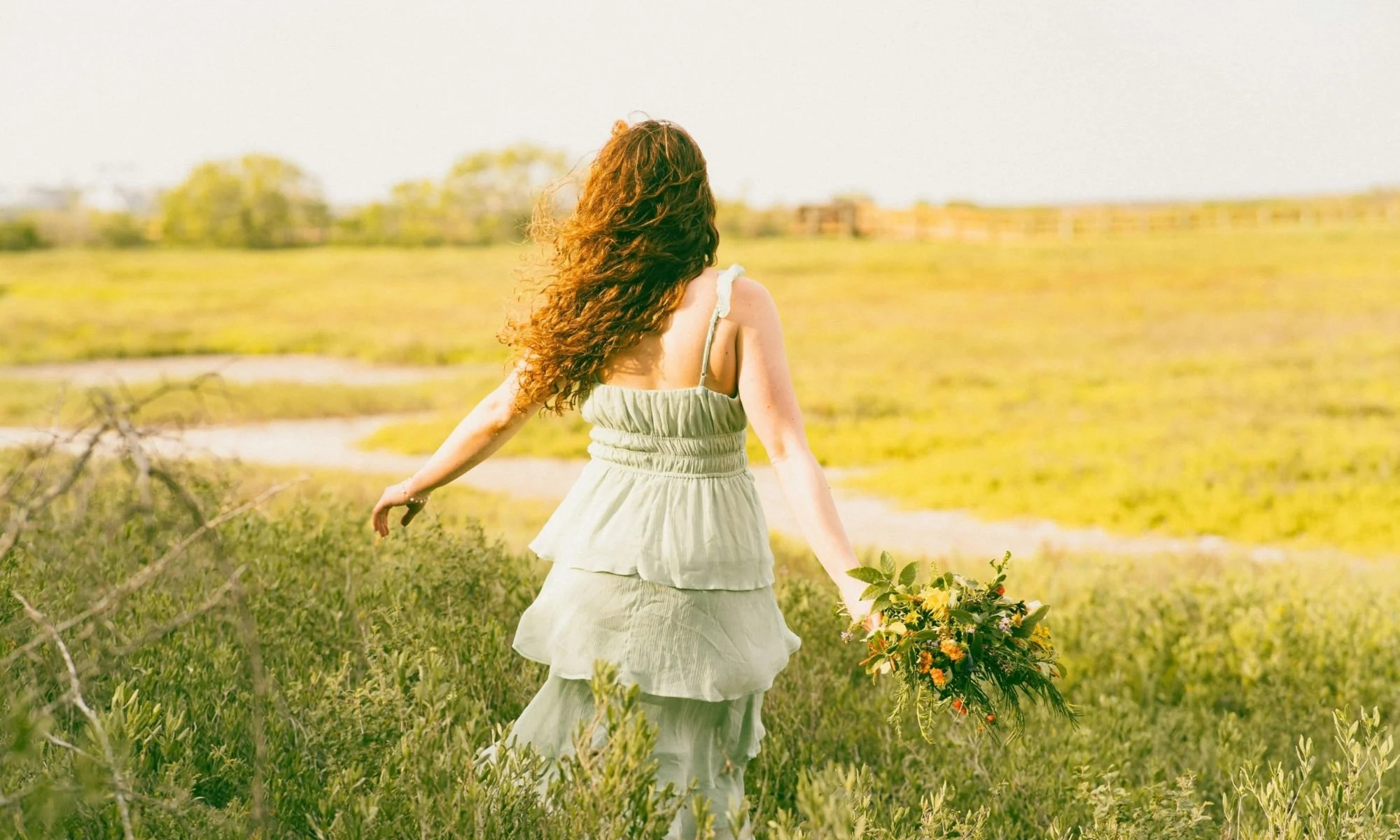 A woman with long, curly red hair wearing a light green sundress walking through a grassy field holding a bouquet of wildflowers.