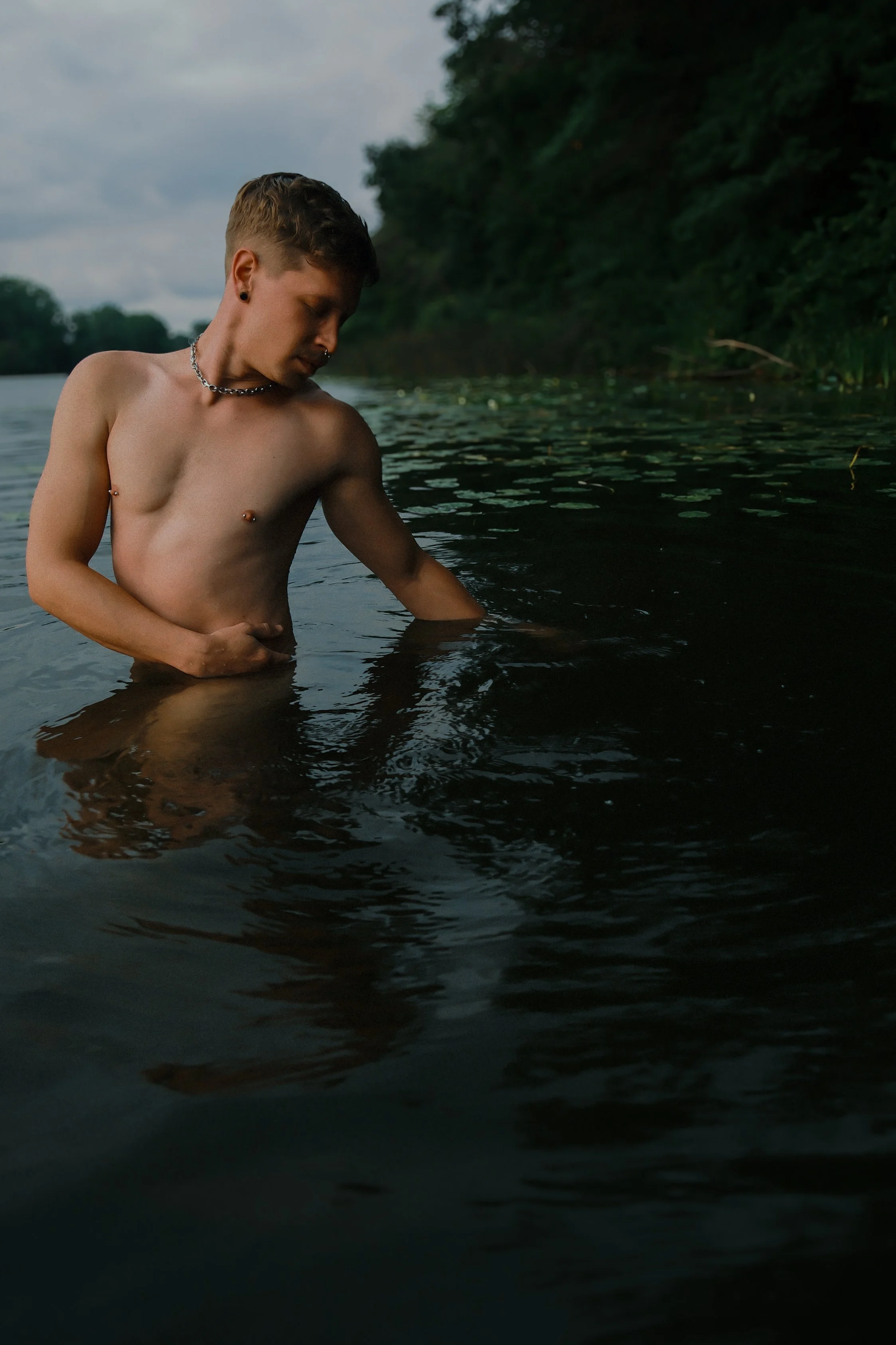 A young person with short hair, earrings, a chain necklace, and a piercing, standing shirtless in a body of water near a forested shoreline, looking down.