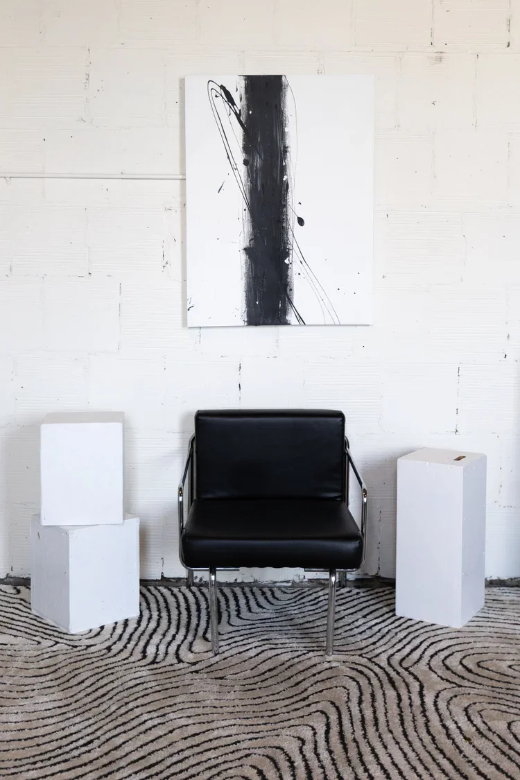 Minimalist interior with a black leather chair, two white pedestal stands, an abstract black and white painting on a white brick wall, and a patterned rug.