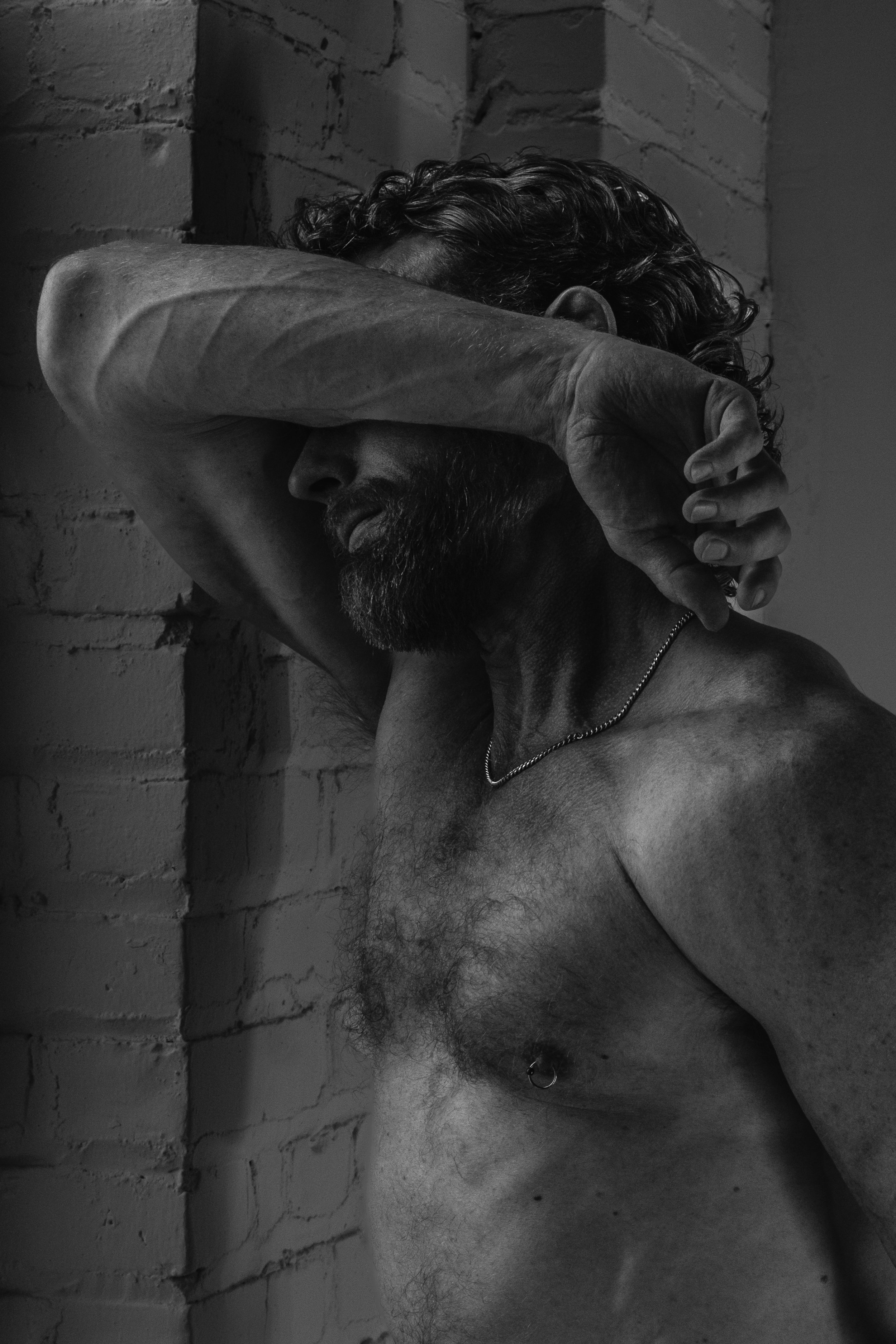A shirtless man with curly hair and a beard, leaning against a brick wall, with one arm covering his face.