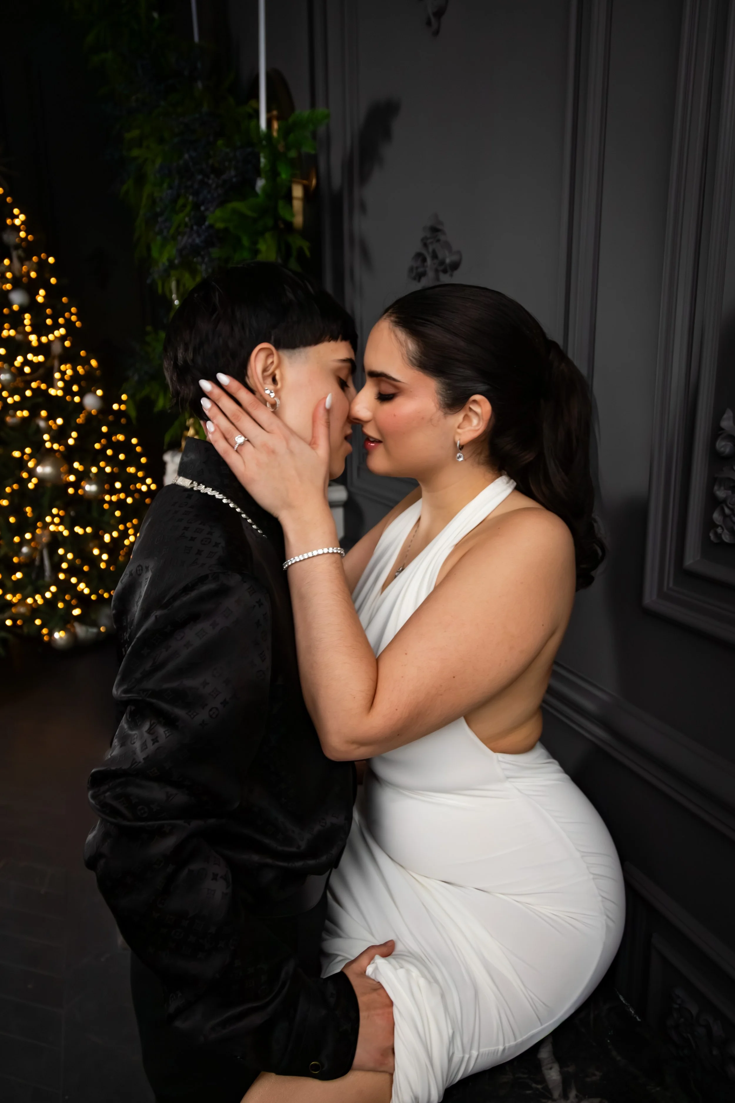 A couple is embracing intimately in a romantic moment, with Christmas decorations and a lit Christmas tree in the background.