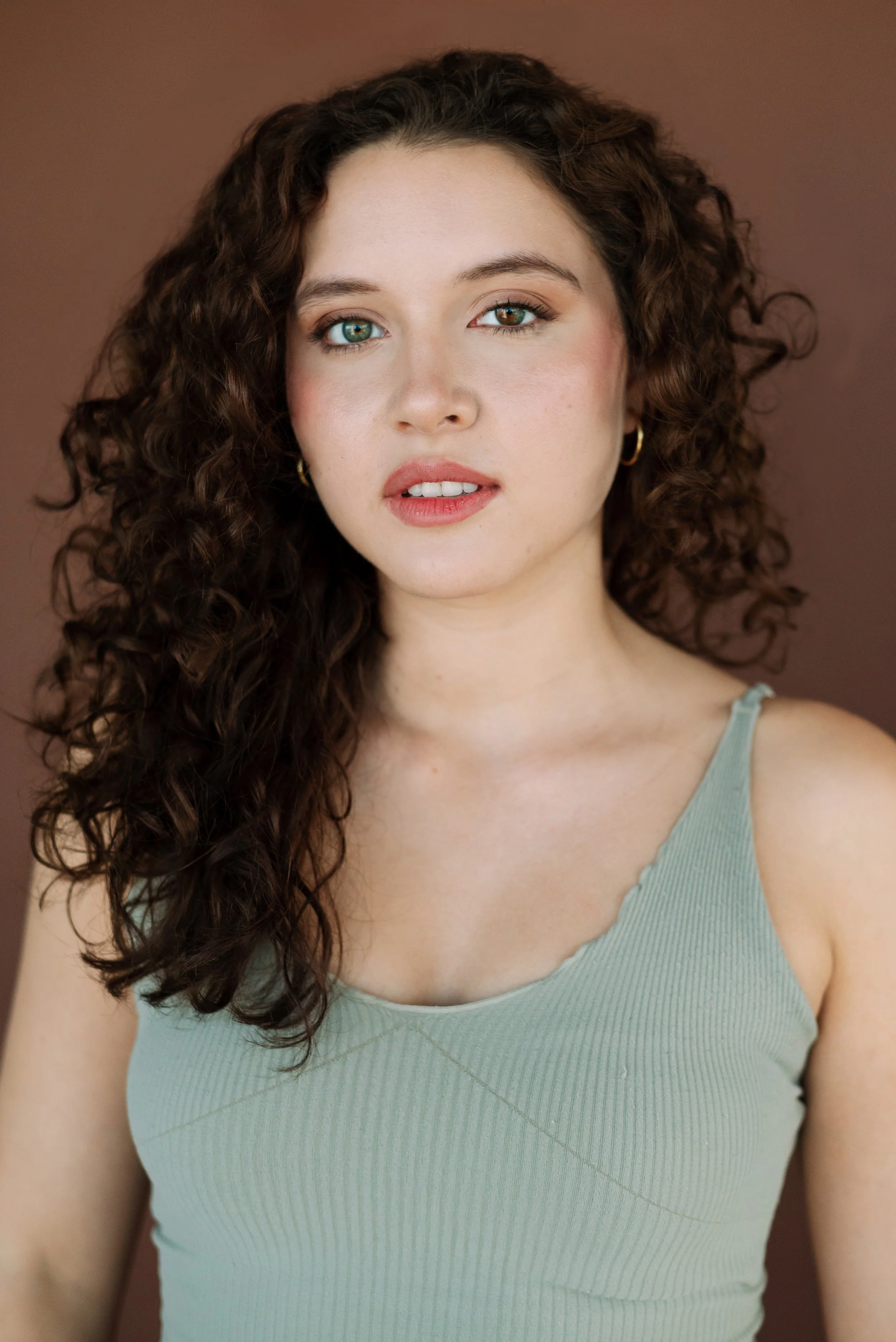 A young woman with curly brown hair, blue eyes, and wearing a light green tank top, posed against a plain brown background.