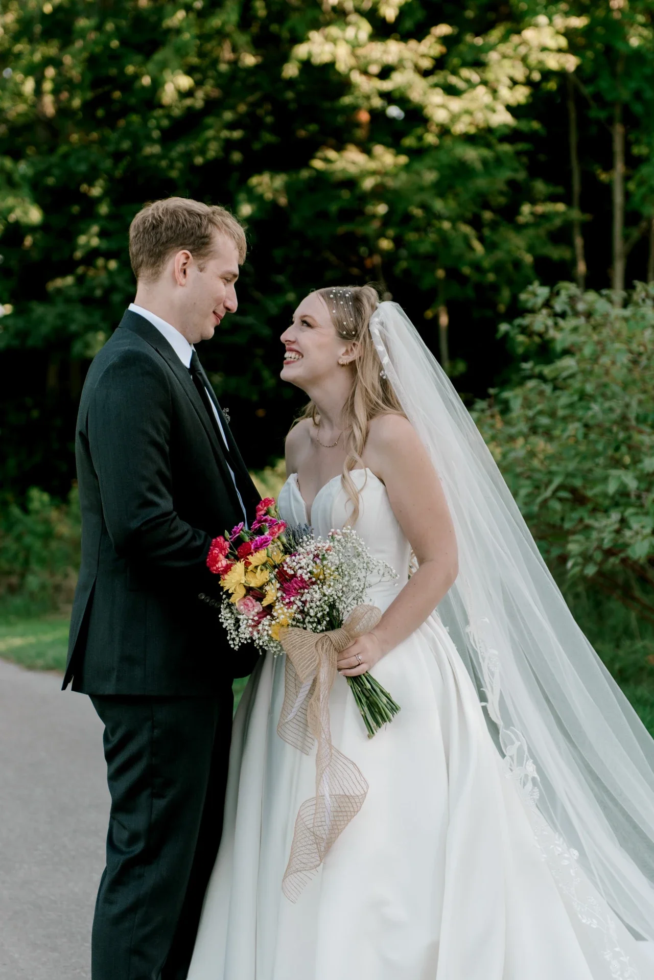 A bride and groom looking at each other and smiling outdoors during their wedding, with the bride holding a bouquet of flowers.
