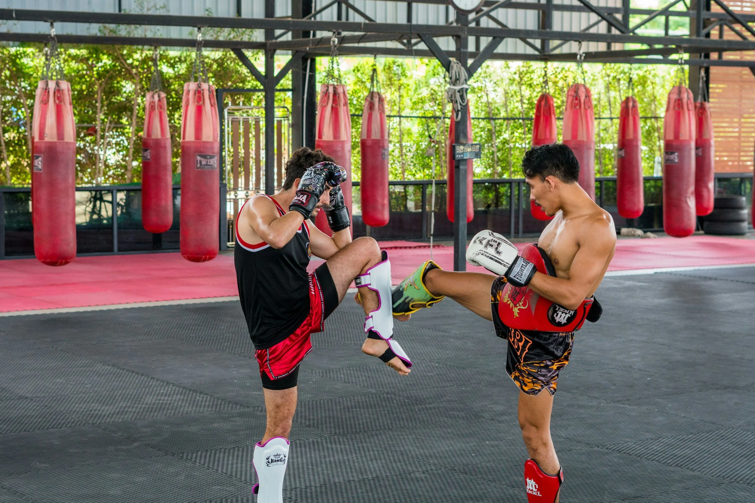 Two shirtless men sparring in a Muay Thai or kickboxing gym, with red punching bags hanging in the background. One man is standing on his left leg with his right leg raised, while the other man is adjusting his gloves or headgear.