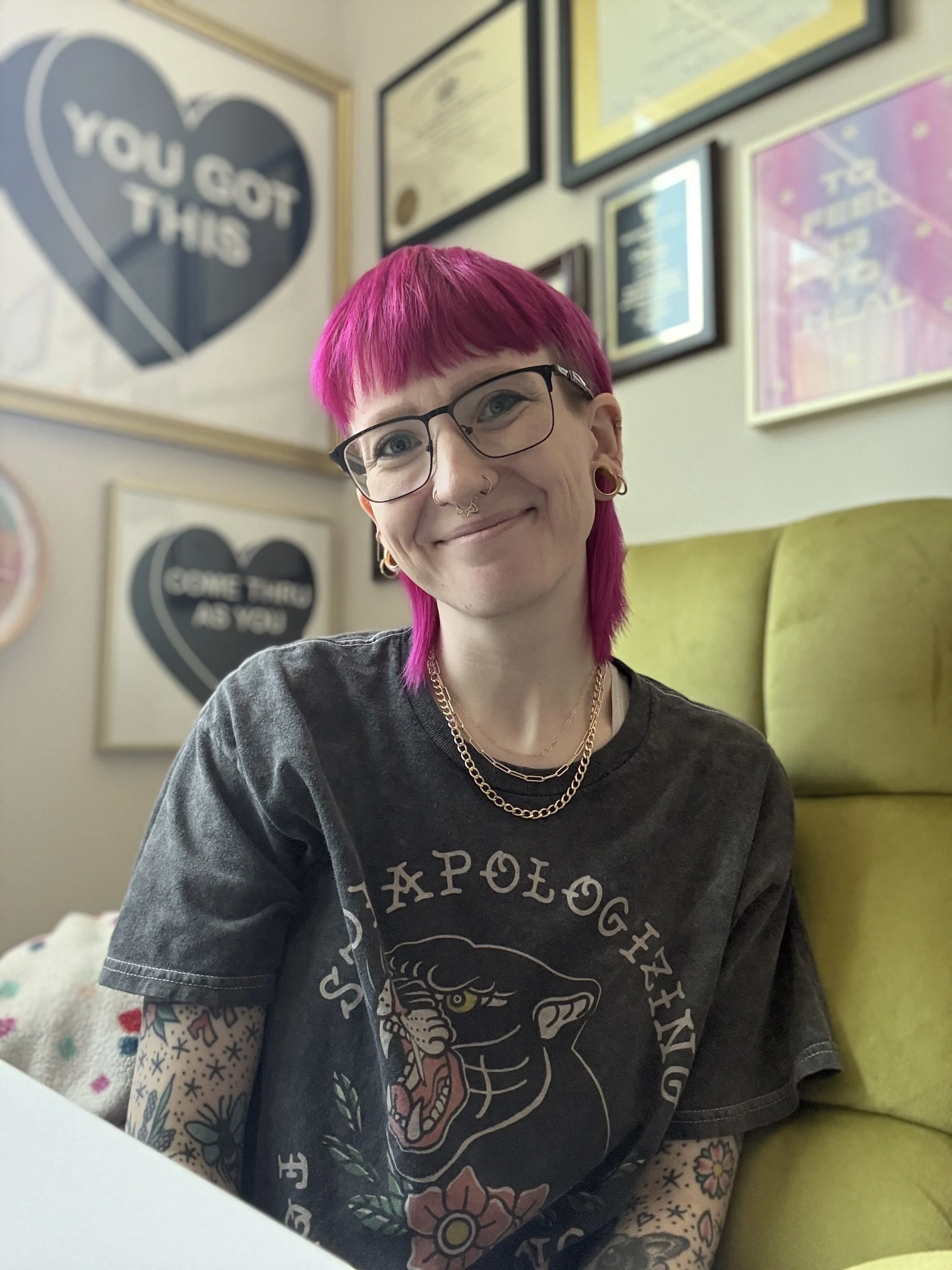 A nonbinary person with vibrant pink hair, glasses, and multiple facial piercings is smiling at the camera. They are wearing a black t-shirt with a tattoo sleeve on their left arm and gold necklaces.