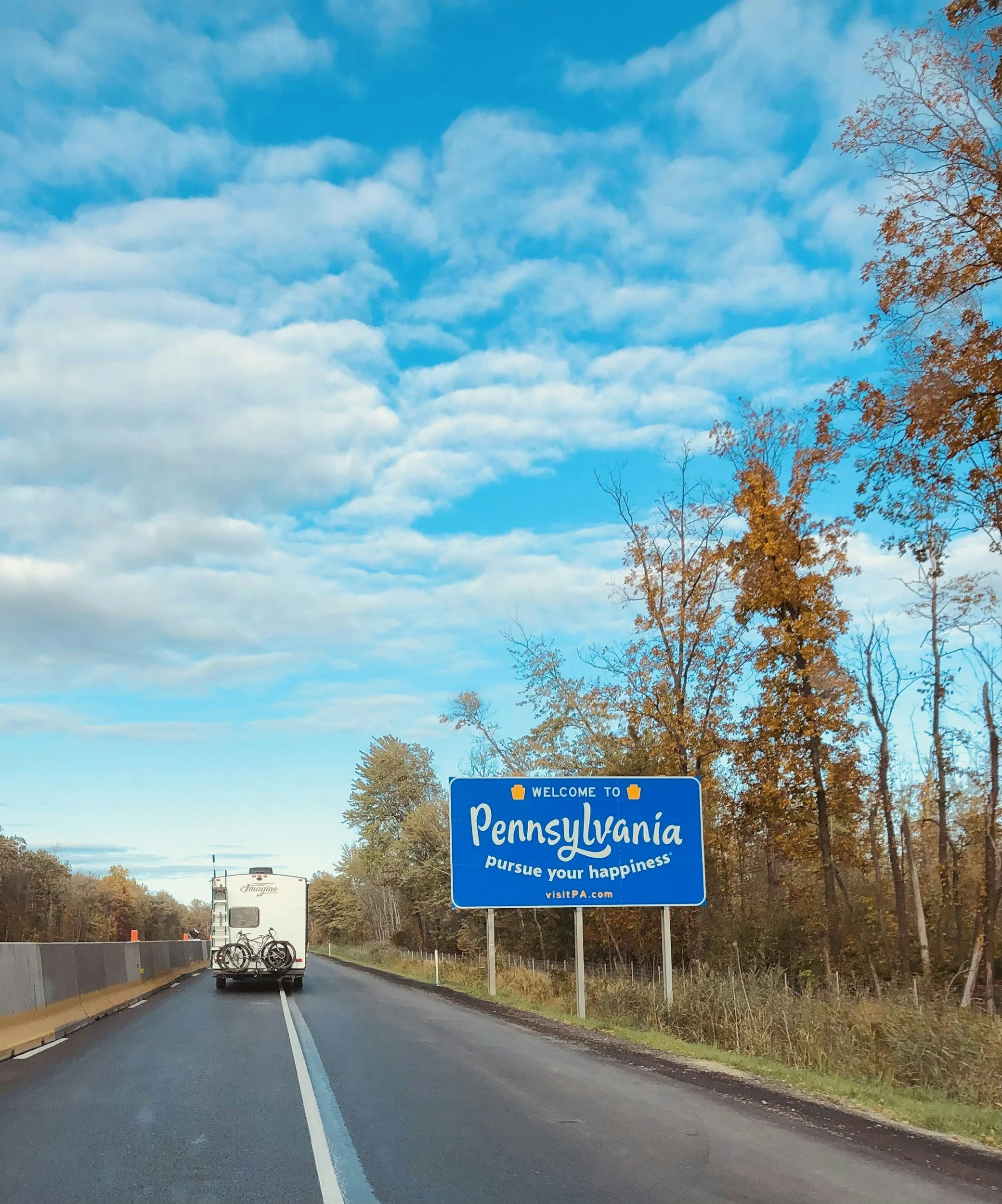 A Blue Pennsylvania state welcome sign on the side of a highway with trees that have orange leaves, indicating an autumn setting. Vehicles are seen driving past the sign.