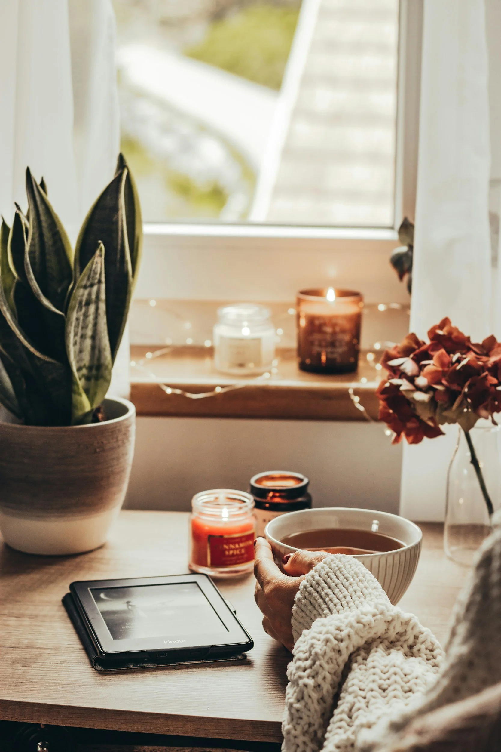 A cozy indoor scene featuring a person's hand holding a cup of coffee or tea, with a wooden table decorated with candles, a potted plant, and a tablet device. The background shows a window with curtains and a view of greenery outside, with warm lighting creating a relaxed atmosphere.