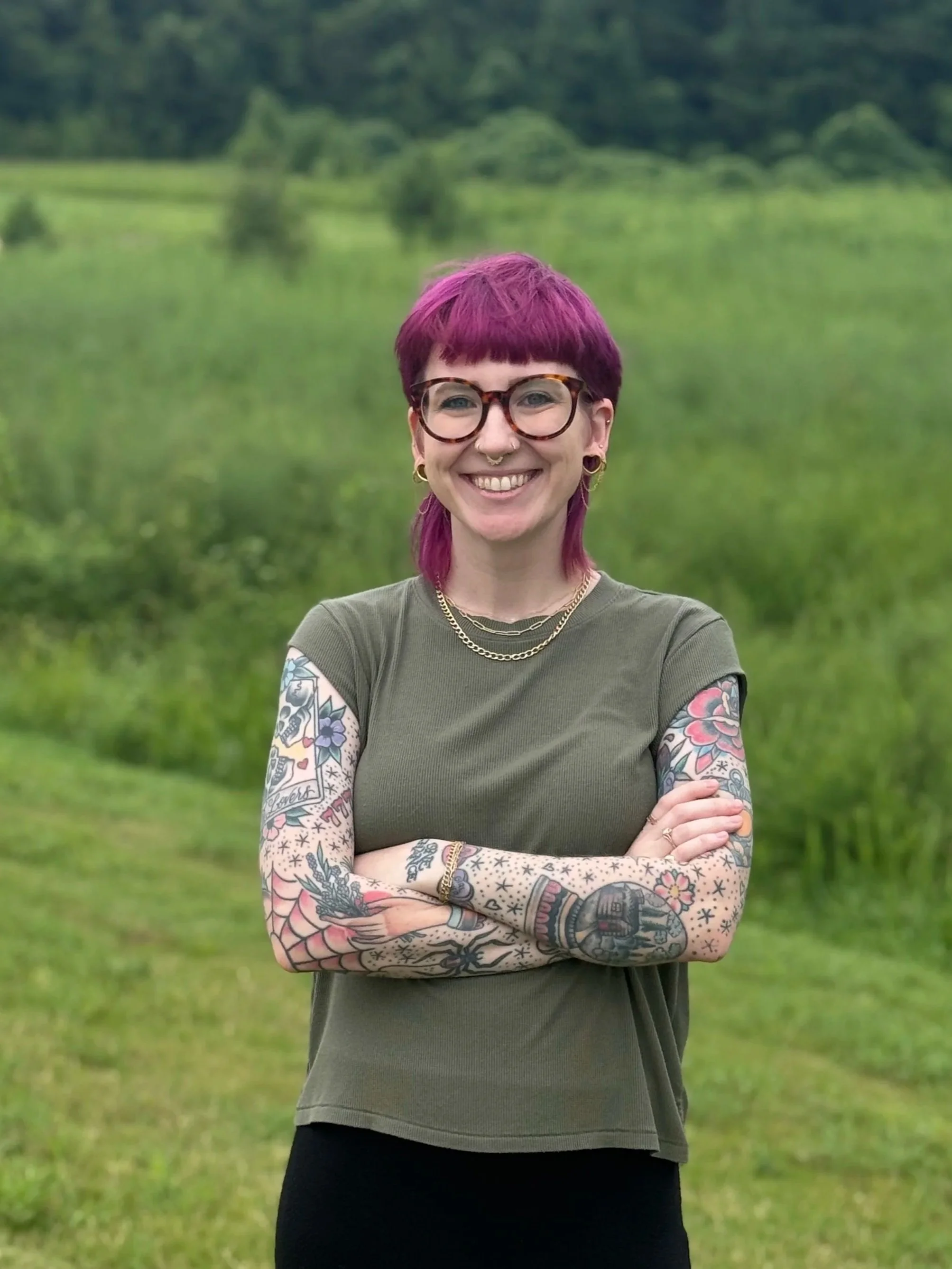 A smiling person with pink hair, glasses, and tattoos on their arms standing outdoors in a green field with trees in the background.