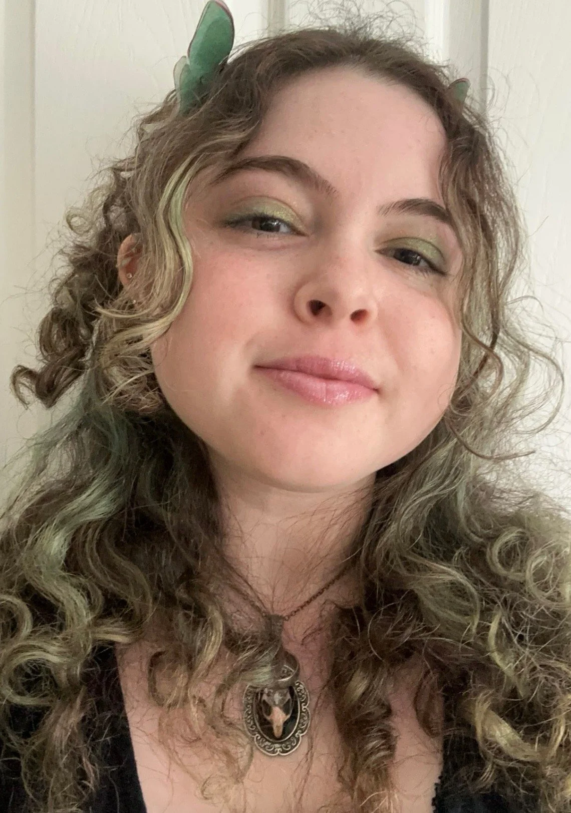 A woman with curly, light brown hair and green eyeshadow is smiling softly, wearing a black top, a necklace with a pendant, and butterfly-shaped hair clips.