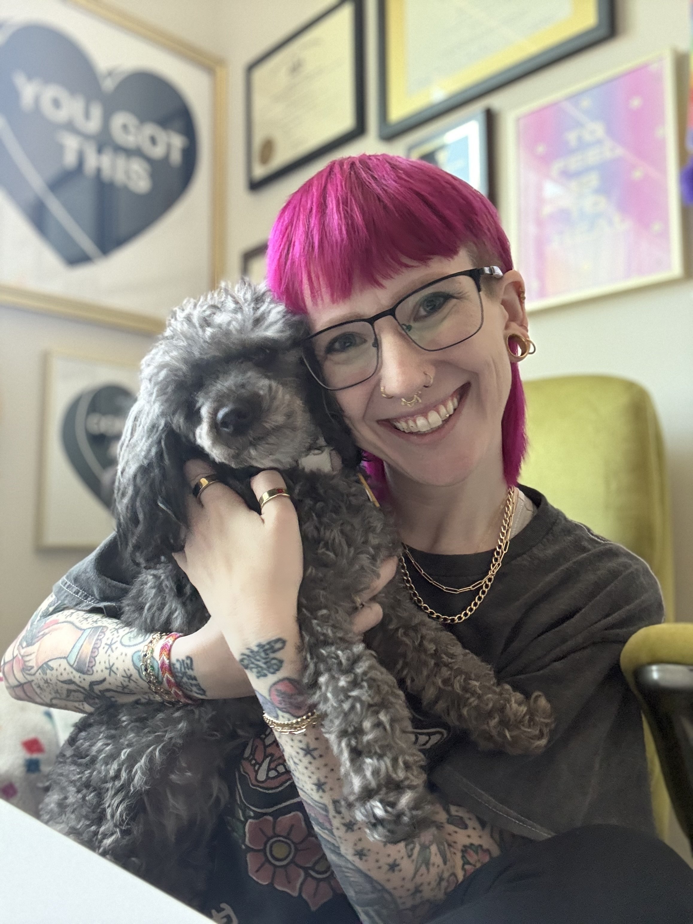 A nonbinary person with bright pink hair, glasses, and tattoos holding a small gray curly-haired dog. The person is smiling widely, showing their teeth, and is wearing gold jewelry. The background shows framed certificates or artwork on the wall.