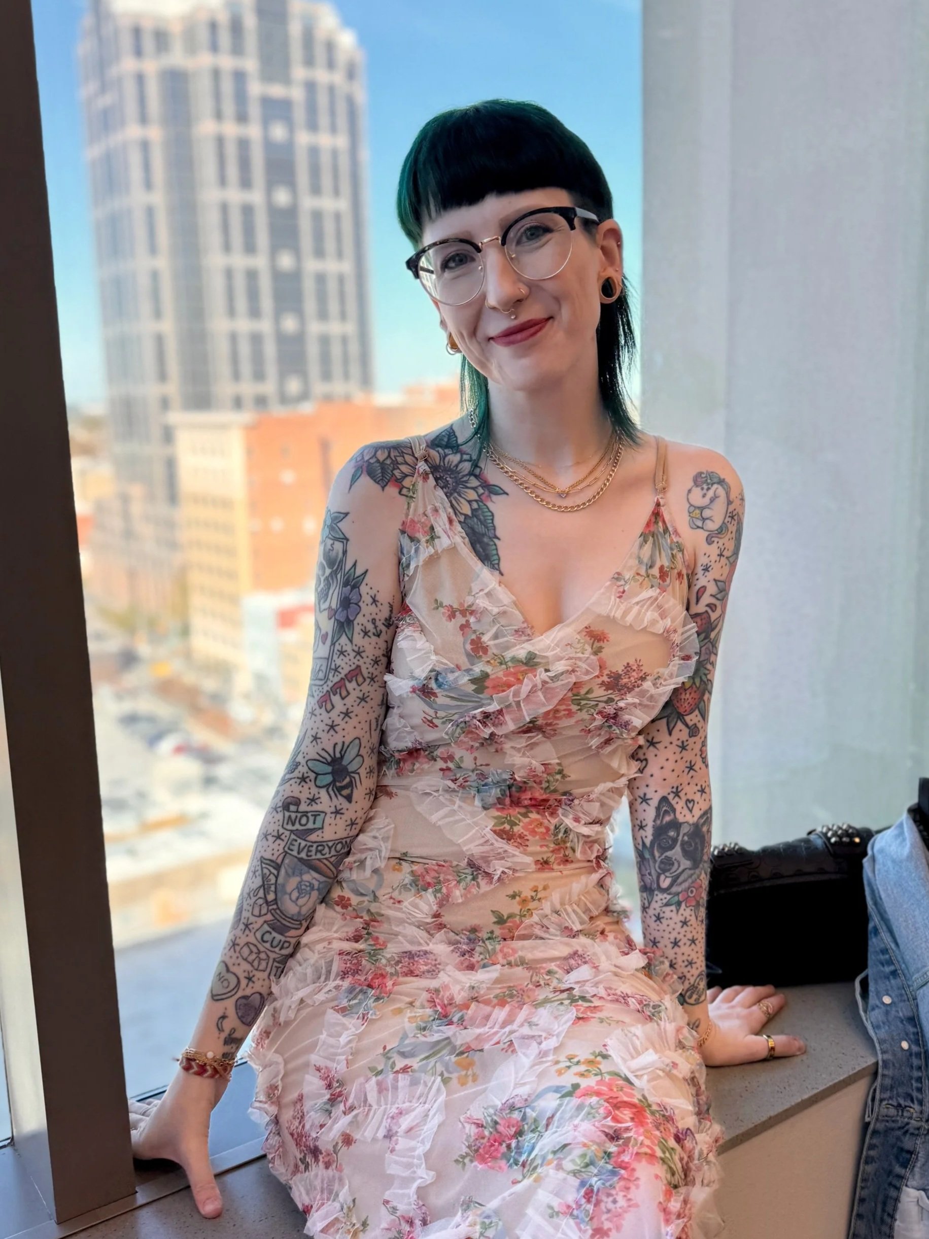 A young person with short dark hair, glasses, and multiple tattoos on their arms, sitting on a windowsill in a city building. They are smiling and wearing a floral, ruffled dress with gold necklaces and earrings.
