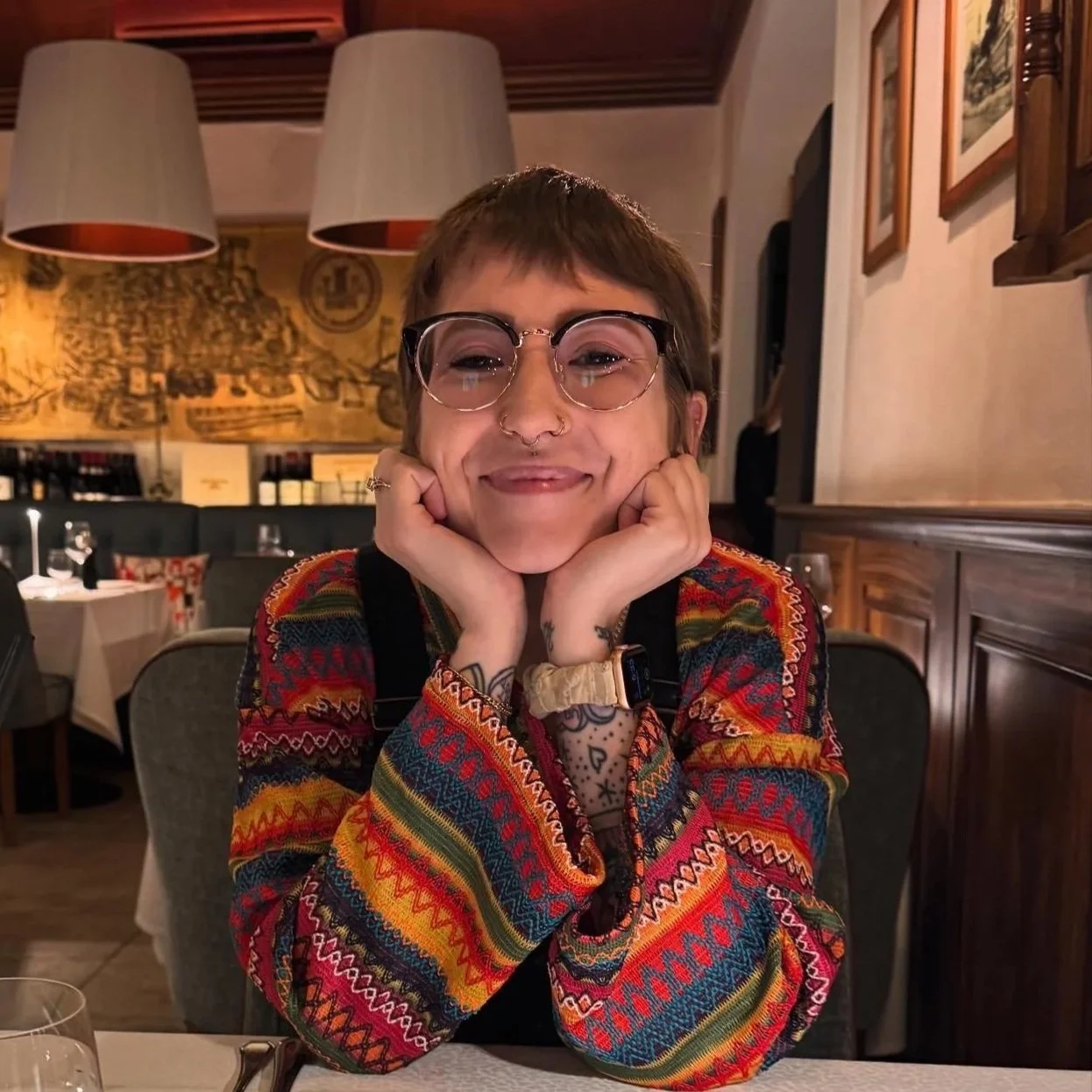 Smiling person with glasses, tattoos, and colorful sweater sitting at a restaurant table with hands resting on face.