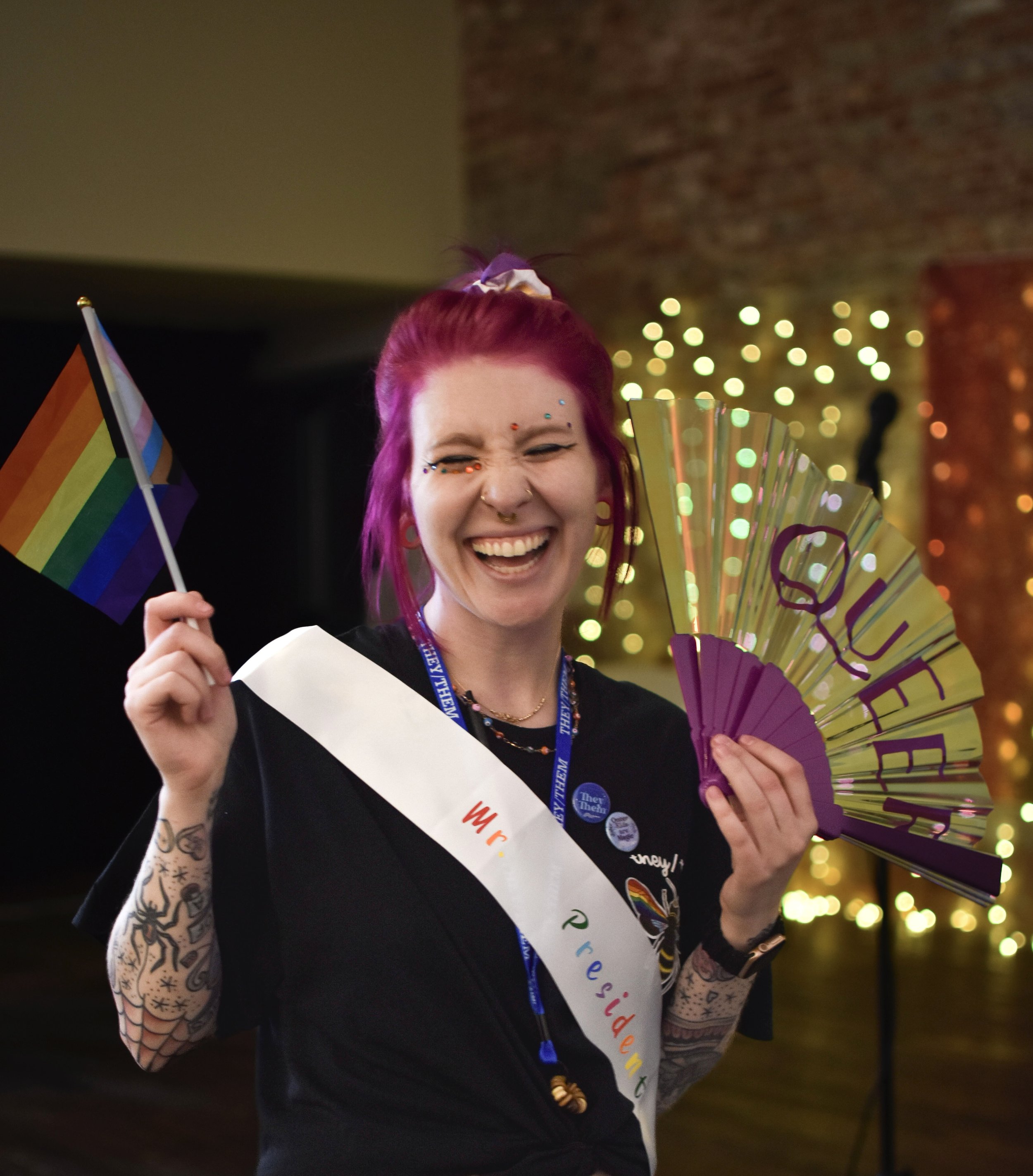 A happy woman with pink hair, wearing a sash that reads 'Ms Pride USA', holding a rainbow pride flag and a rainbow-colored fan, smiling with closed eyes, in a celebration setting with colorful lights in the background.