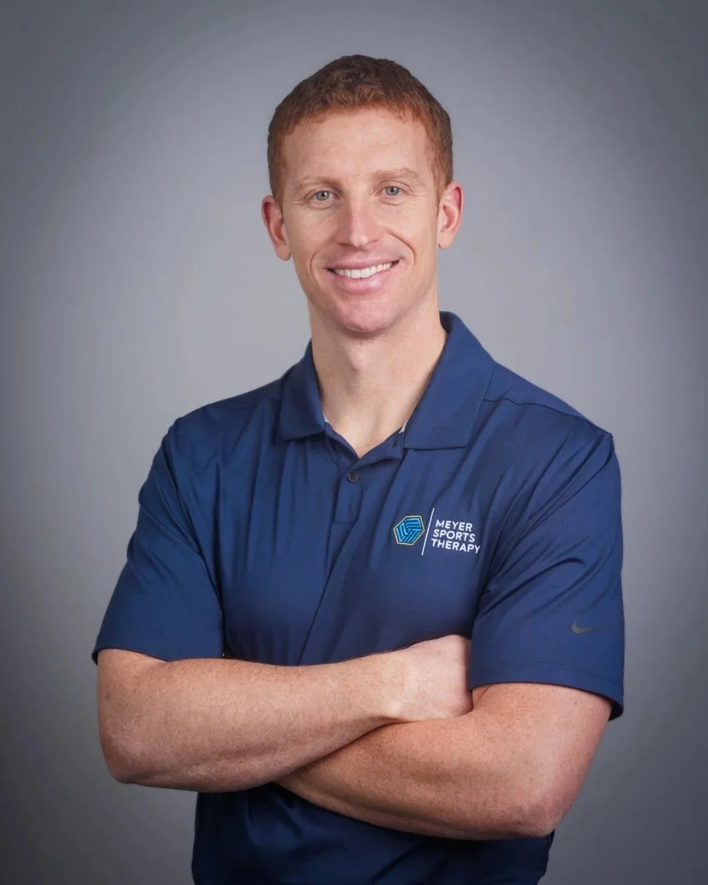 A man with short red hair and fair skin wearing a navy blue polo shirt with 'Meyer Sports Therapy' logo on the chest, smiling with crossed arms against a gray background.