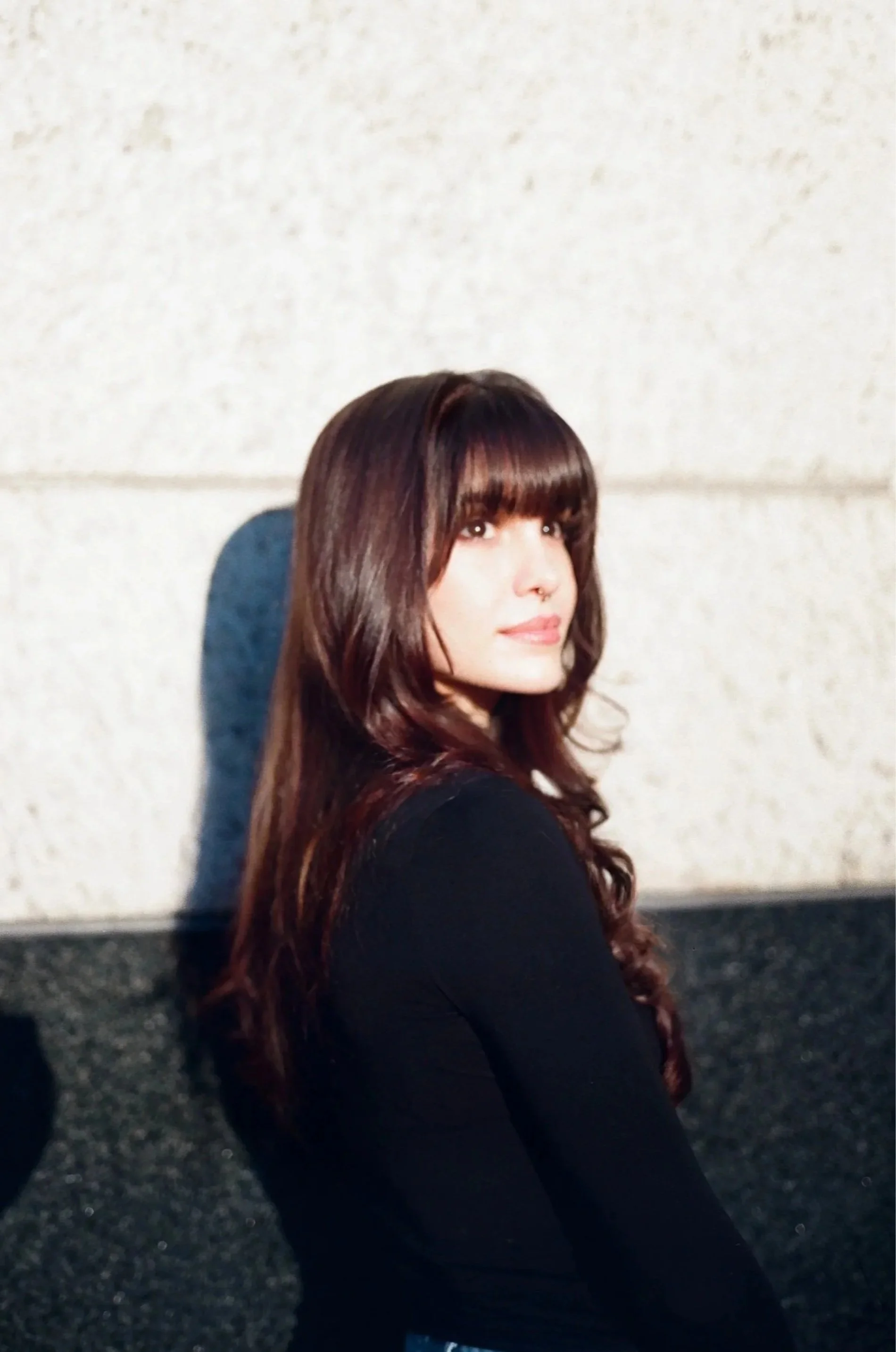 A woman with long, wavy brown hair and bangs, wearing a black outfit, is sitting on a dark, gravelly ground with her back against a light-colored wall, looking slightly over her shoulder towards the camera.