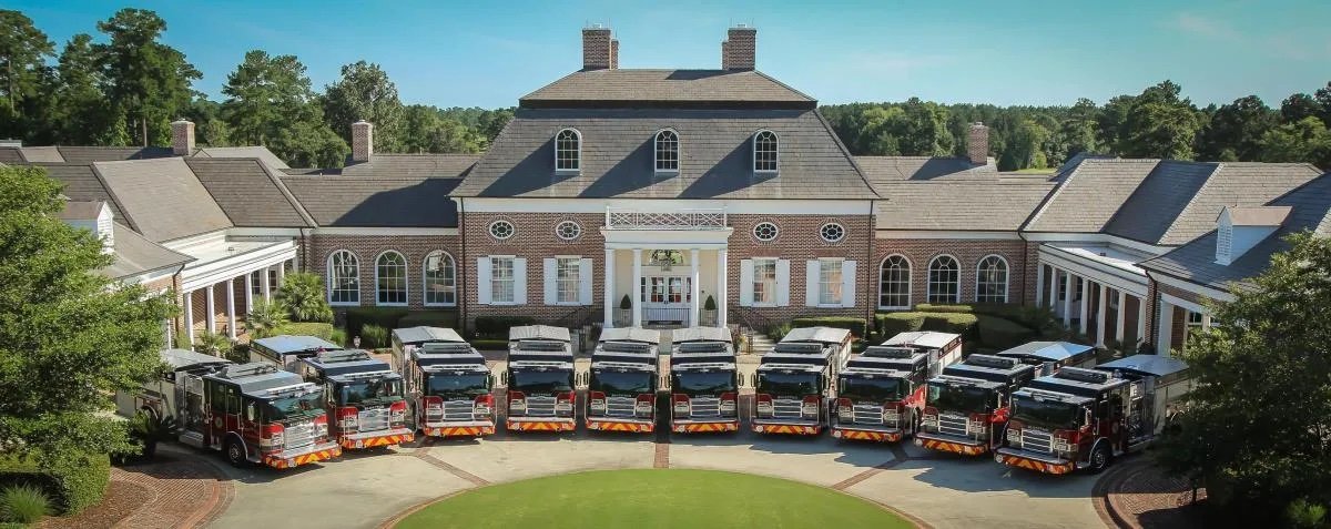 Multiple fire trucks parked in front of a large brick building with a circular driveway and green lawn