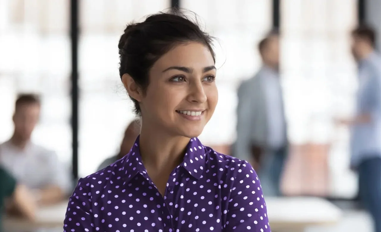 A woman with short dark hair and a bright smile, wearing a purple blouse with white polka dots, in a bright office with blurred people in the background.