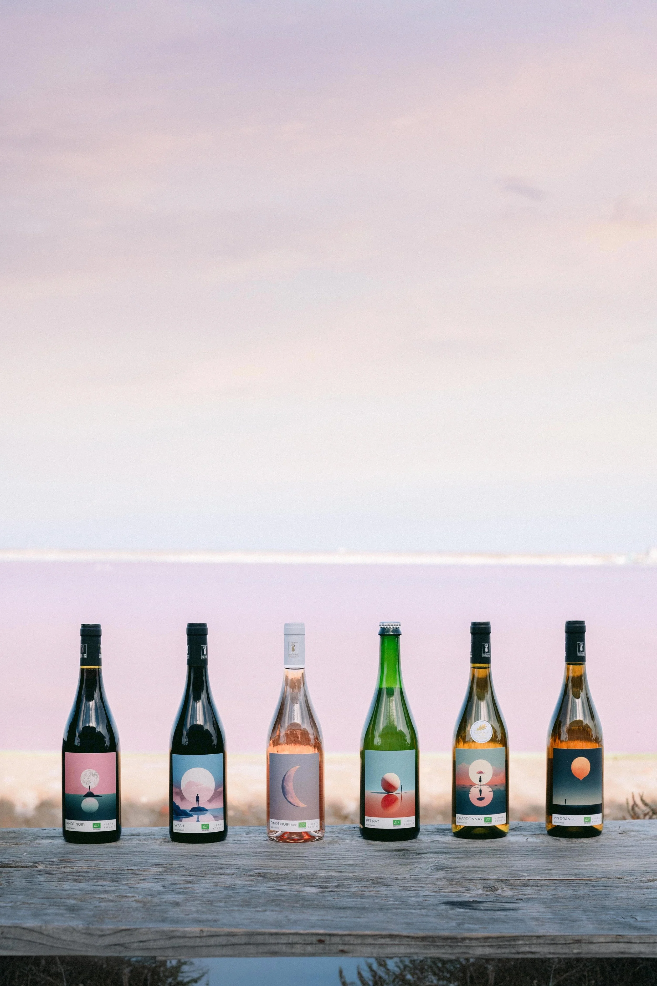 Six bouteilles de vin aux étiquettes artistiques alignées sur une table en bois, avec un paysage de coucher de soleil en arrière-plan.
