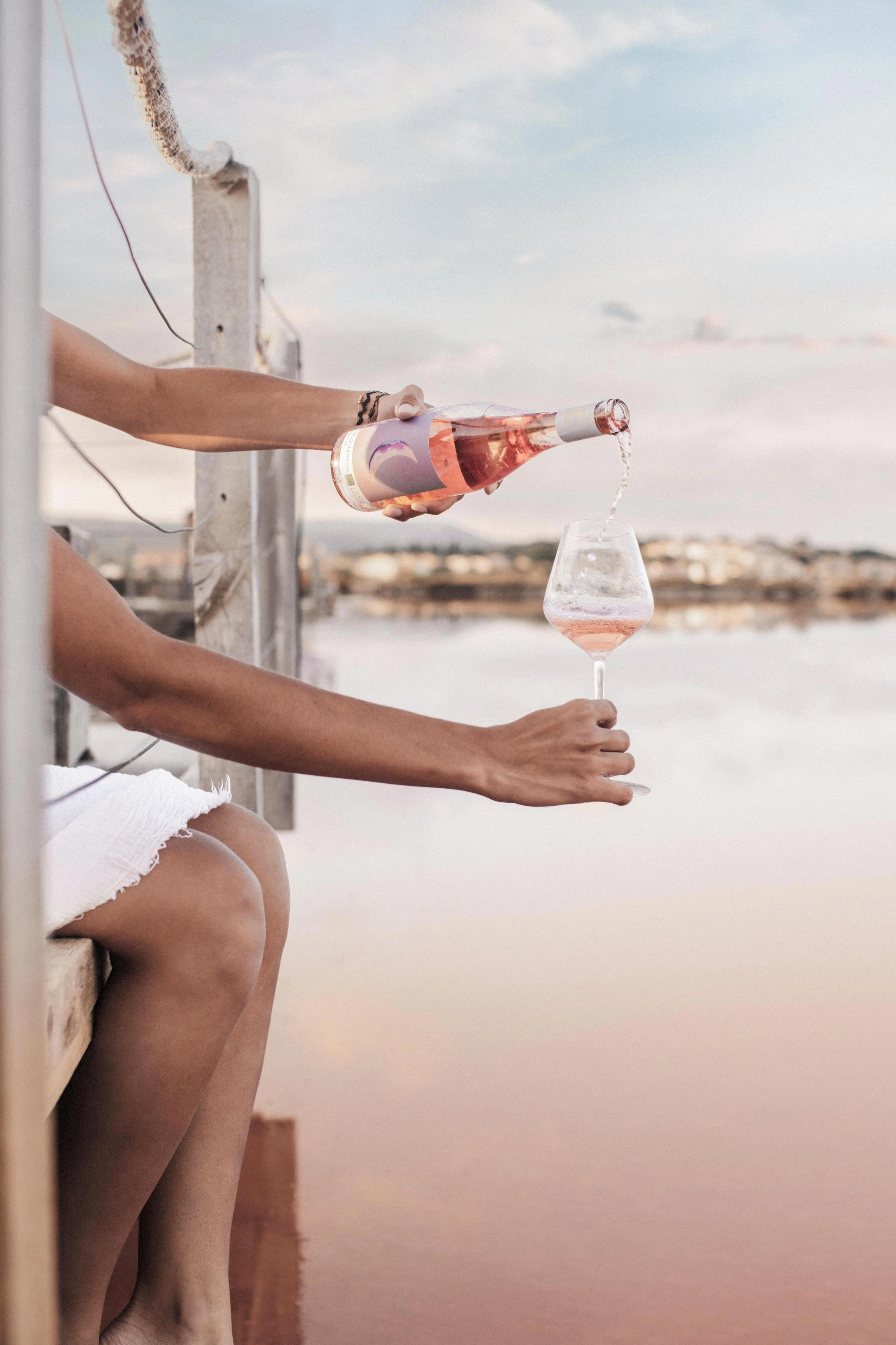 Une personne versant du vin rosé dans un verre au bord de l'eau.